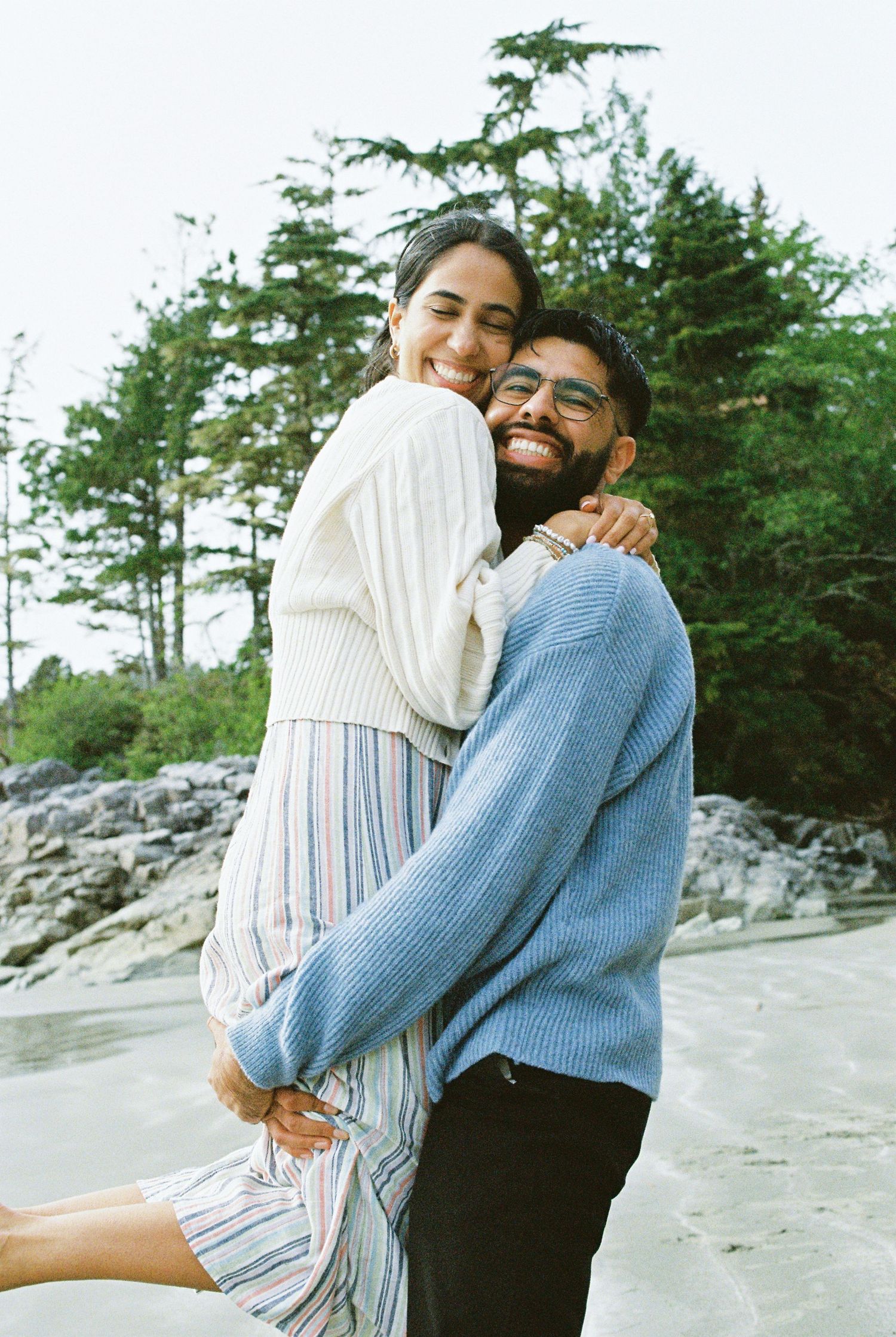 Joyful couple laughing and embracing on a beach with evergreen trees behind them.