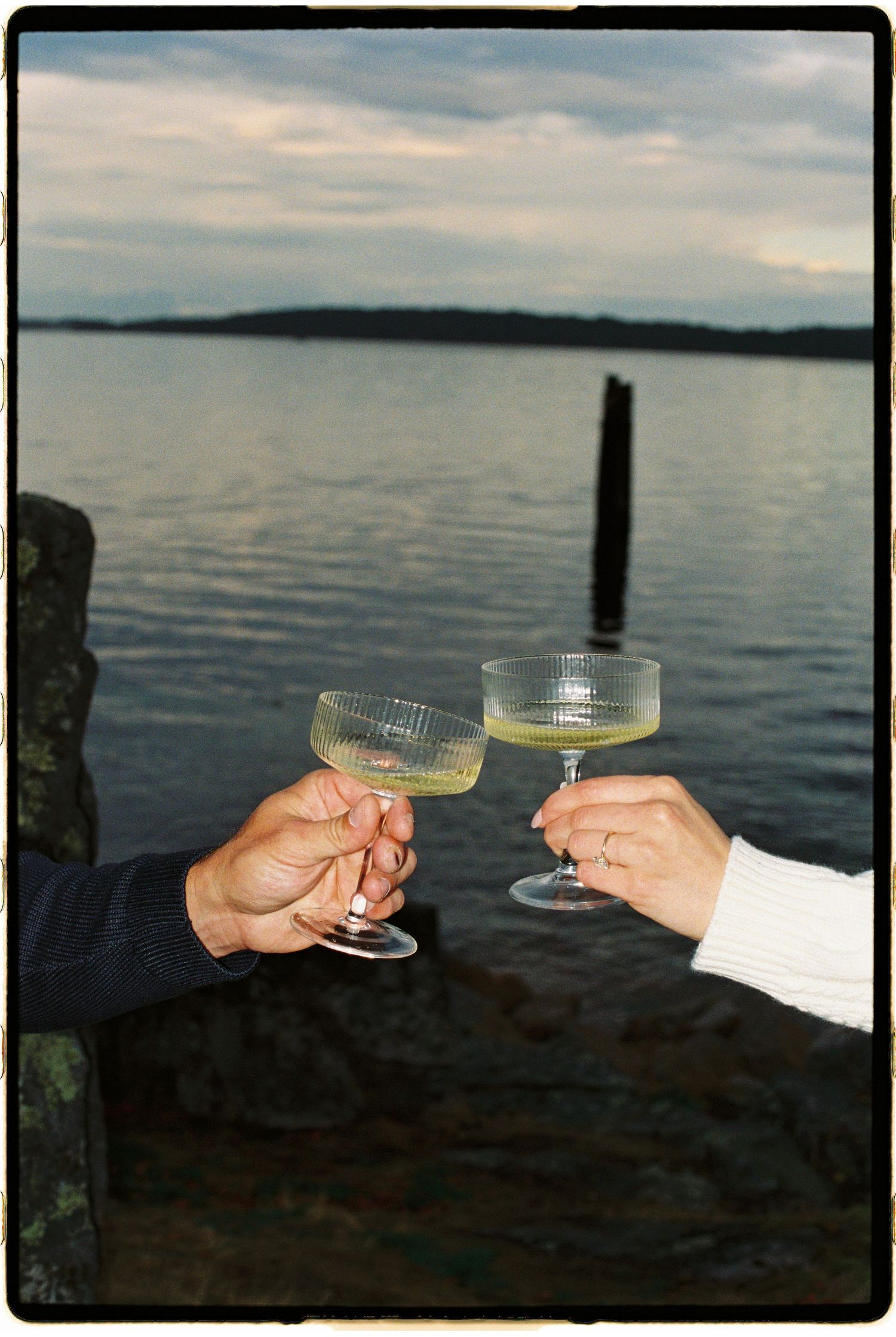 Two people toasting with elegant coupe glasses filled with yellow cocktails near a serene lake.