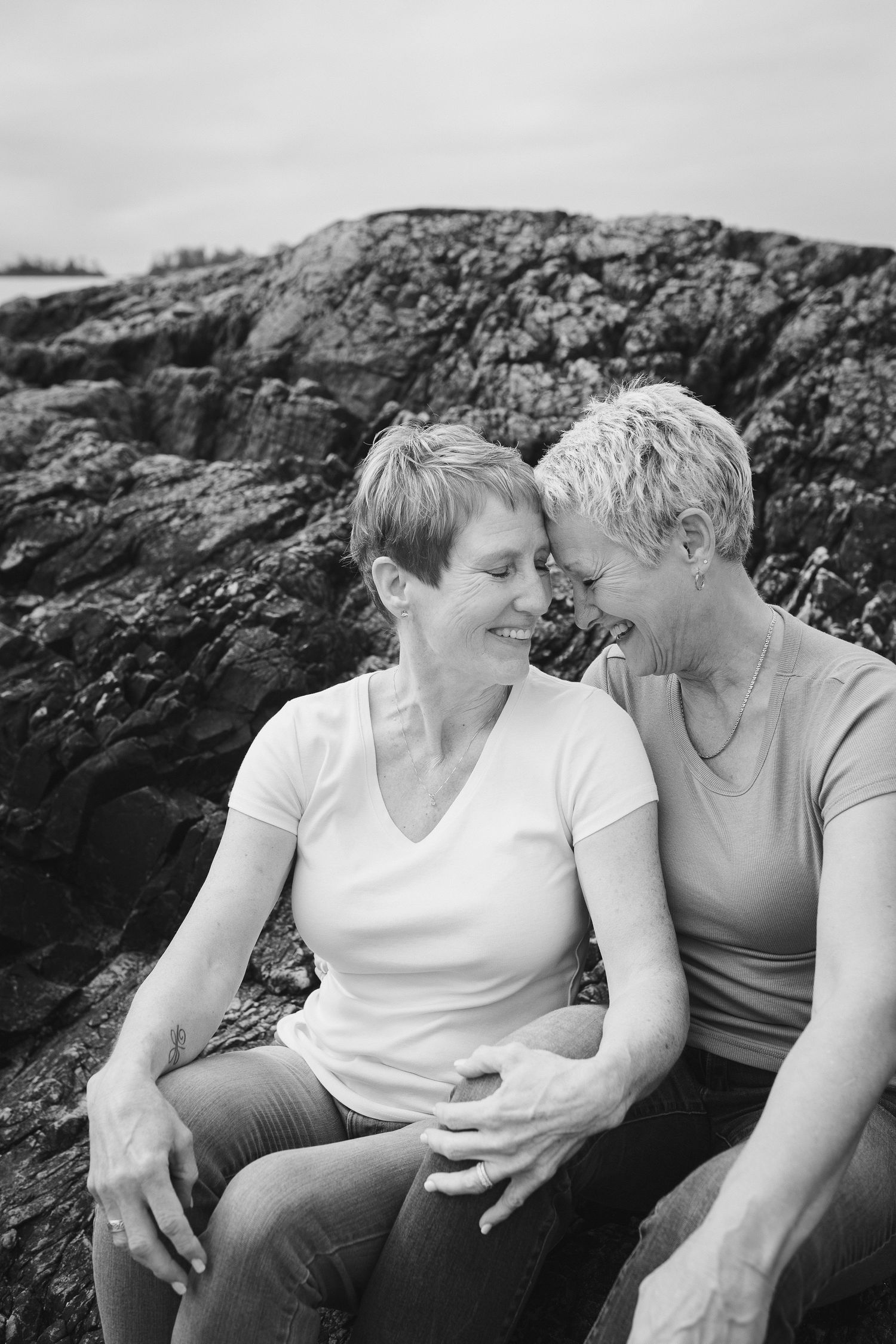 Black and white photo of two women laughing together while sitting on rocky coastal terrain.