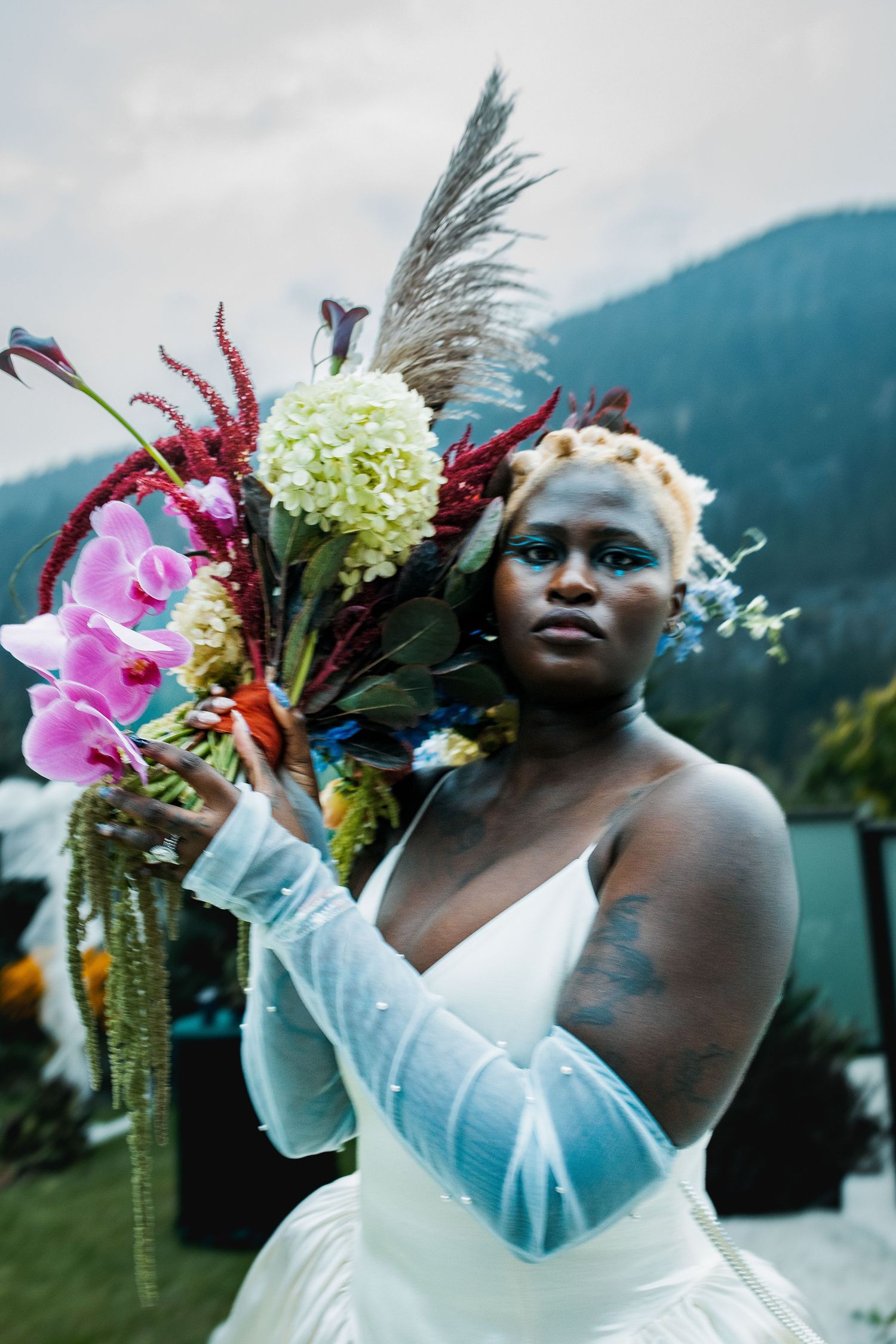 Black bride in white gown holds vibrant bouquet with orchids and pampas grass against mountain backdrop.