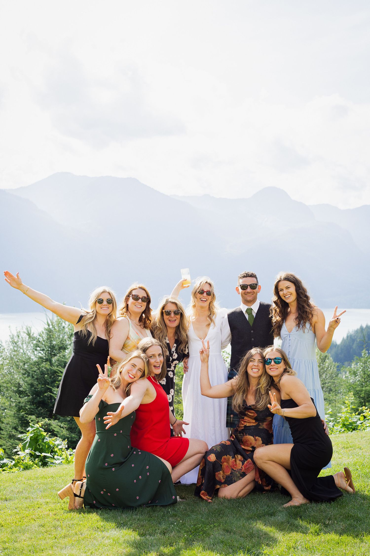 Joyful wedding guests in colorful dresses posing with peace signs on scenic mountain overlook.