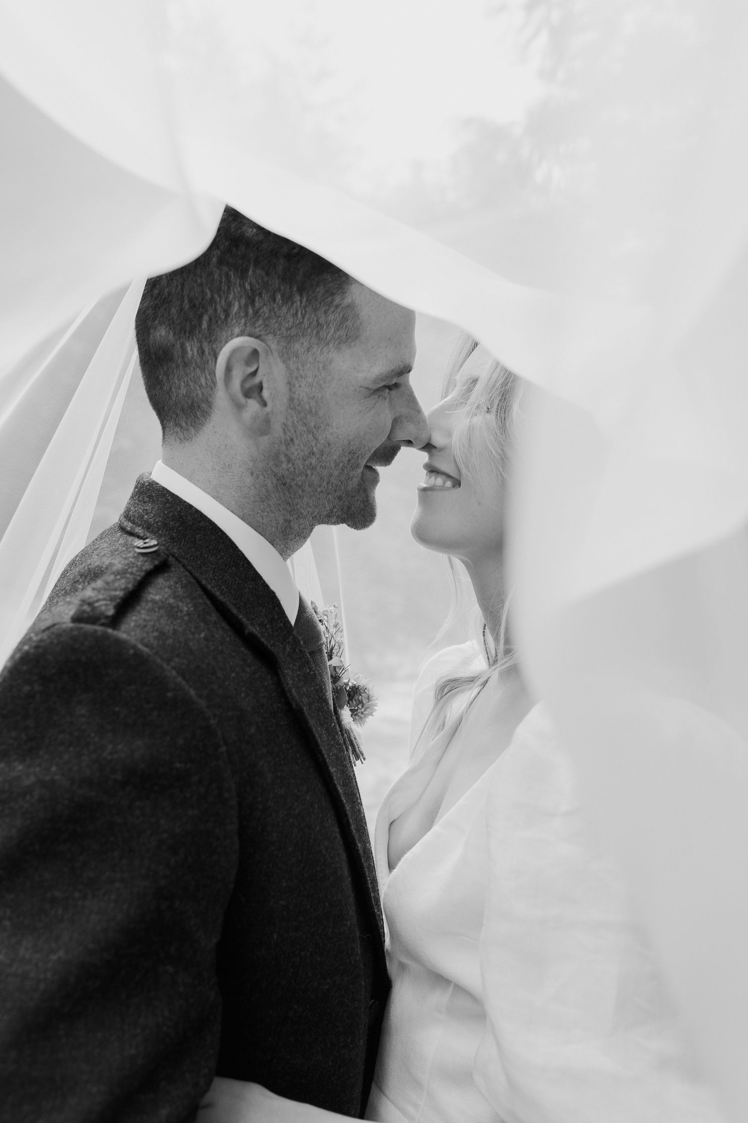Black and white photo of smiling bride and groom under flowing veil together.