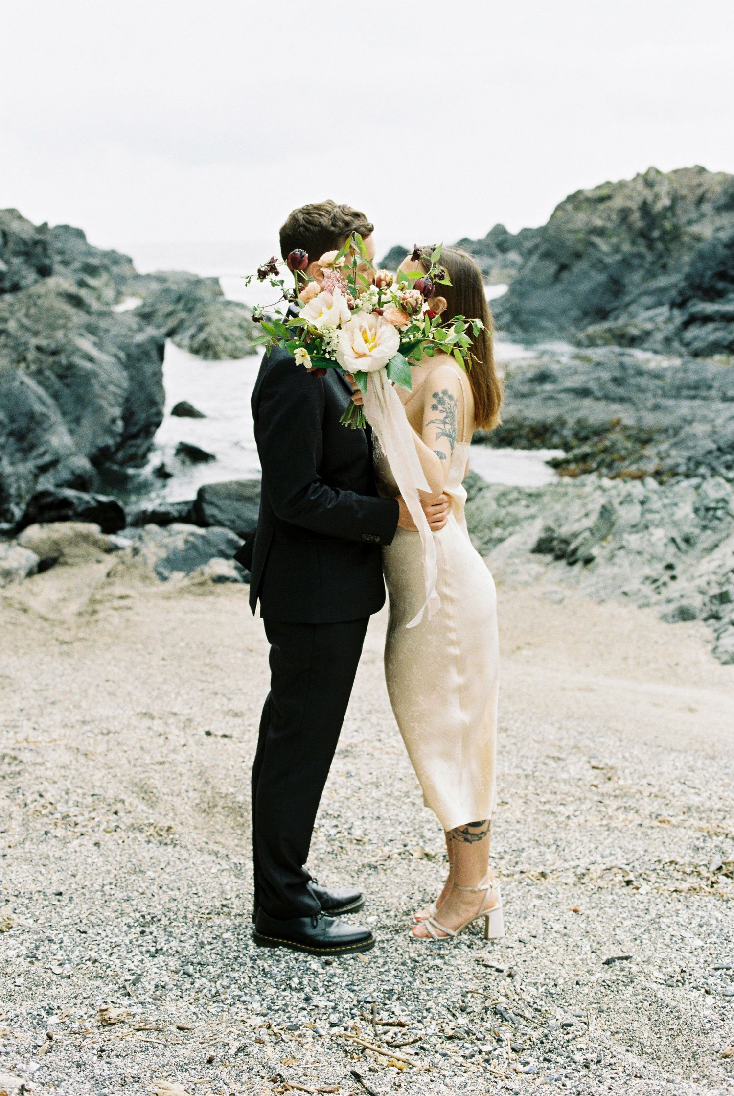 Couple kissing behind a lush floral bouquet on a rocky coastal beach at their elopement.