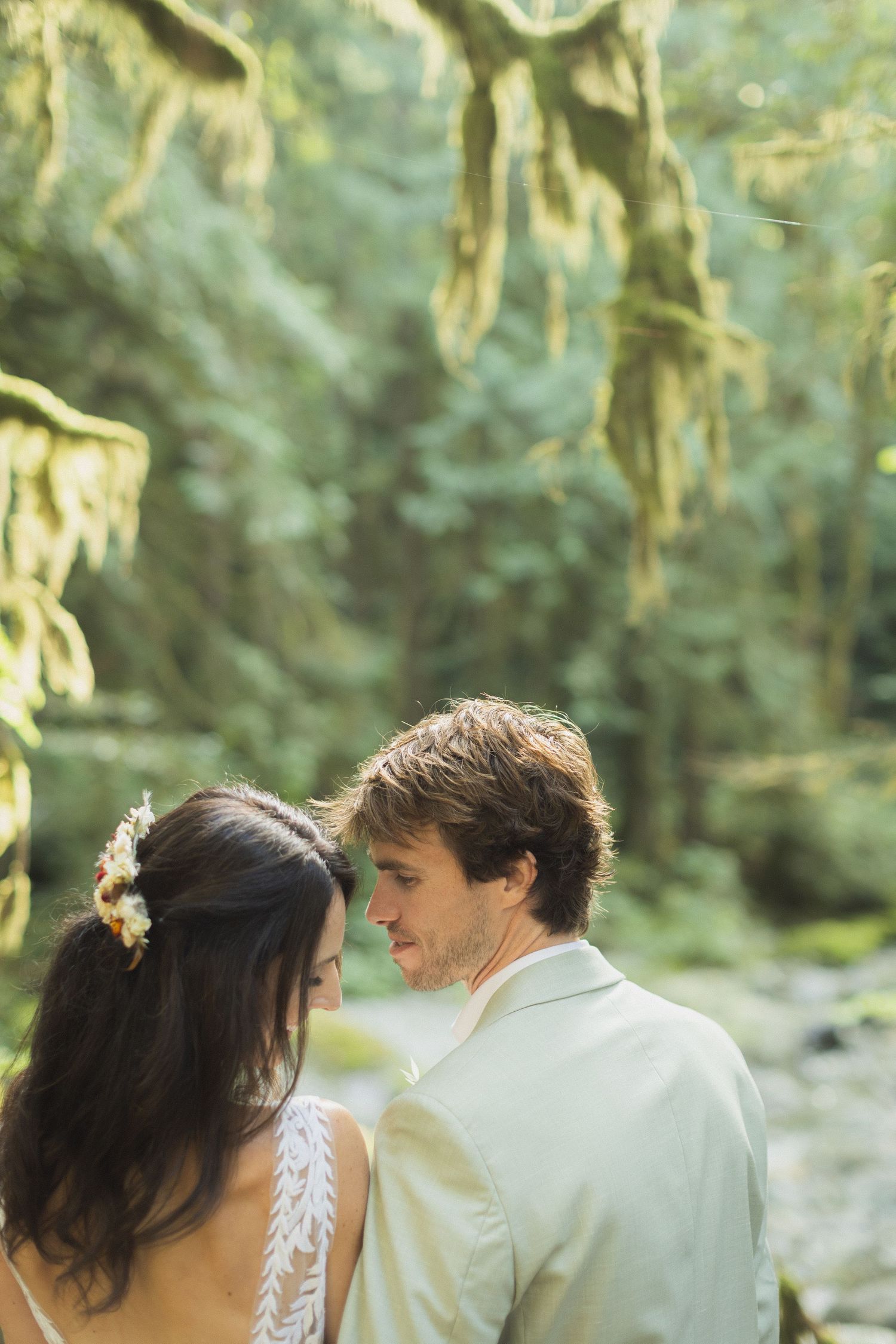 Bride and groom share intimate moment surrounded by lush green moss-draped forest trees.