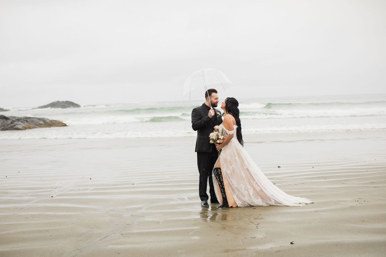Newlyweds share a kiss under a clear umbrella on a foggy beach after their elopement ceremony.