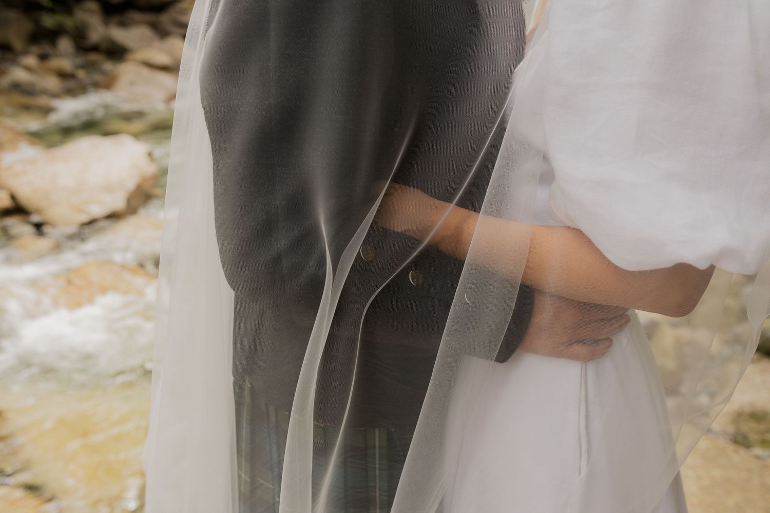 Close-up of bride and groom embracing, draped in a flowing wedding veil by a rocky stream.