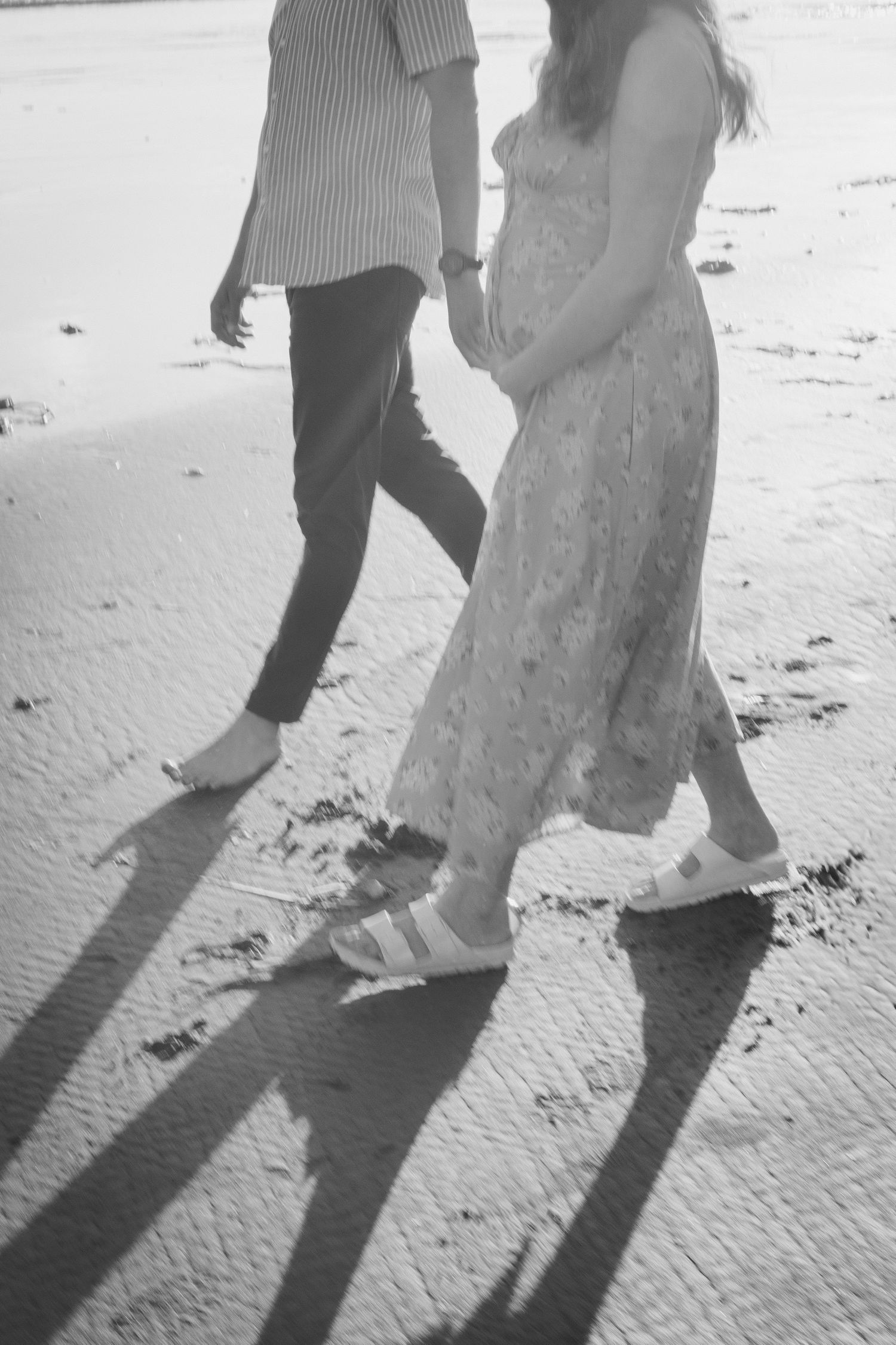 Black and white photo of pregnant couple walking barefoot on sandy beach, casting long shadows in sunlight.