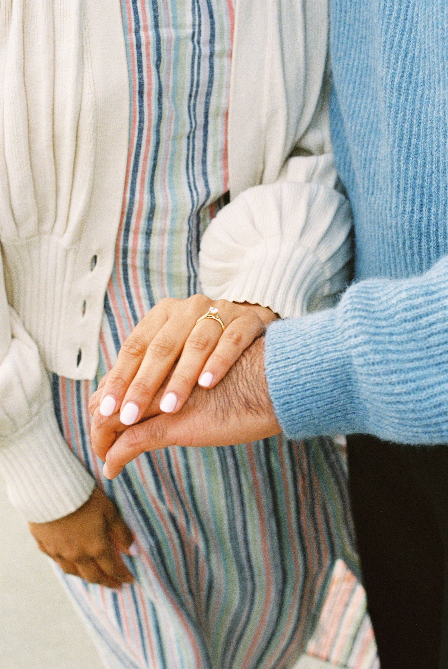 Close-up of couple holding hands, woman wearing white nail polish and delicate gold ring, dressed in striped and white outfits.