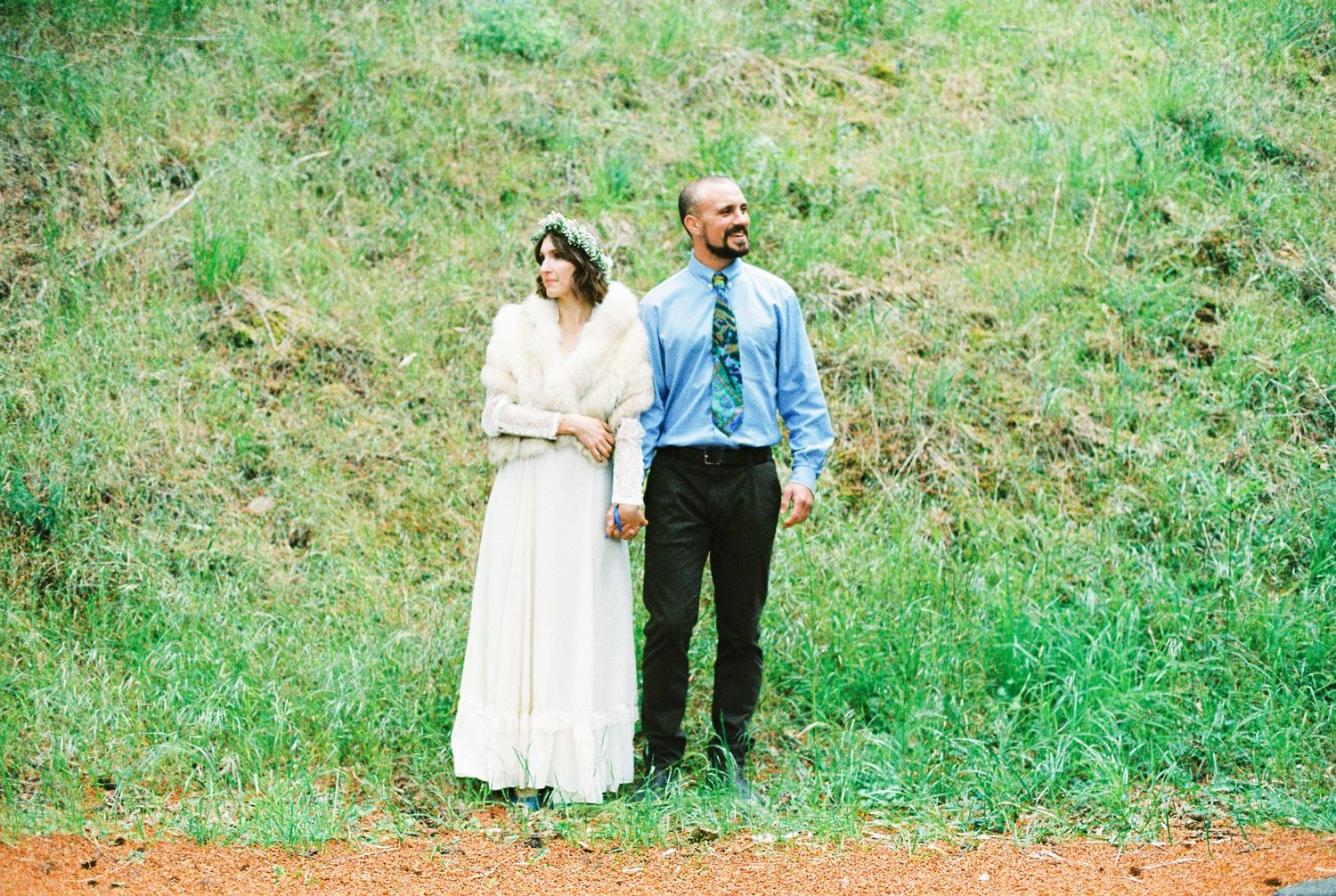 Bride in vintage white dress and groom in blue shirt holding hands on a grassy hillside.