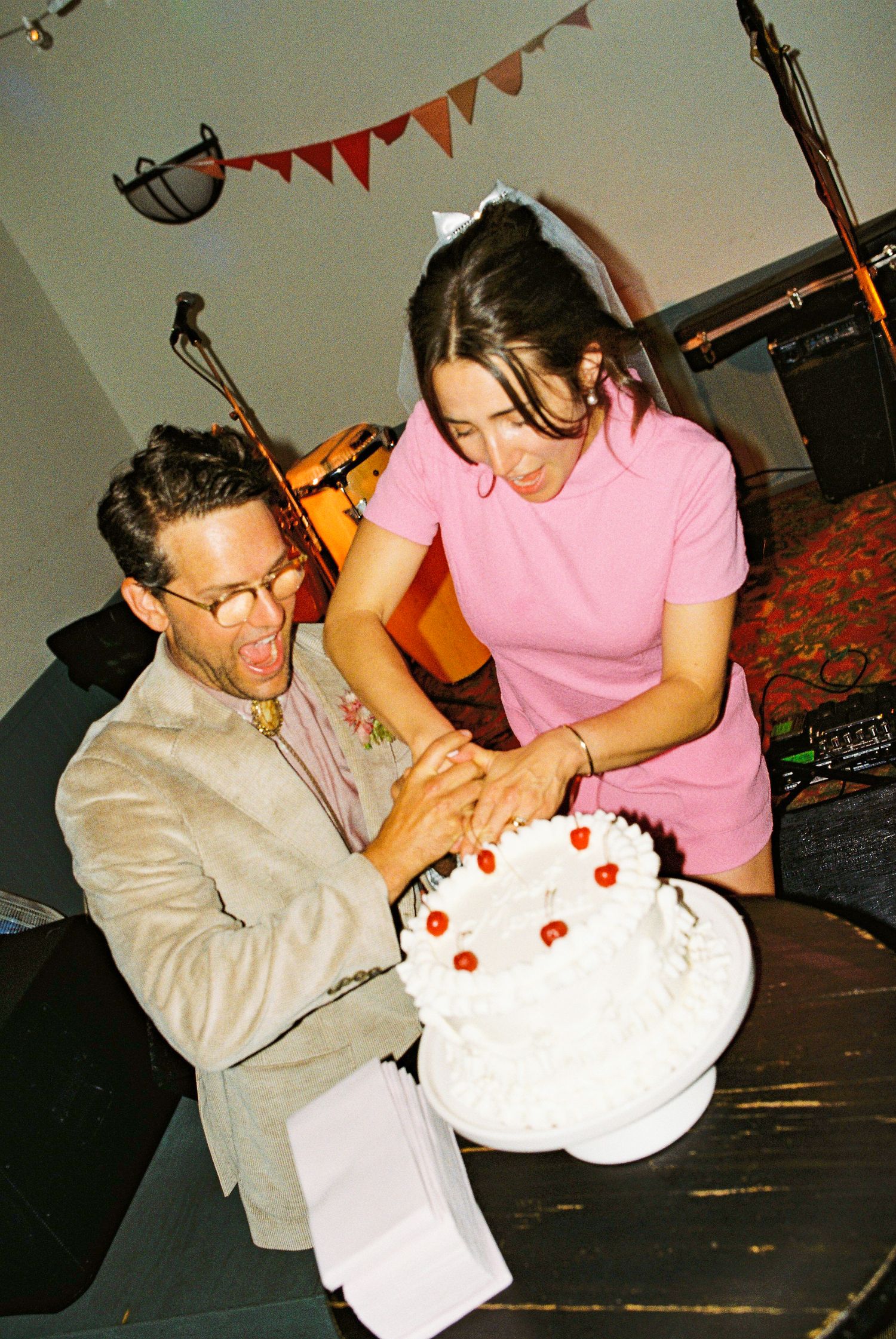 Groom in linen suit playfully opens mouth as bride in pink dress cuts white wedding cake with cherries.
