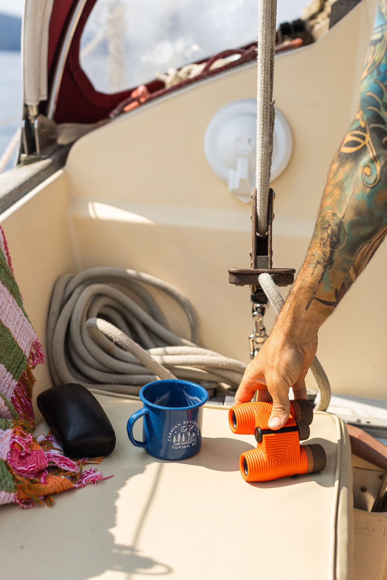 Tattooed hand picking up orange binoculars on sailboat deck with blue enamel mug nearby.