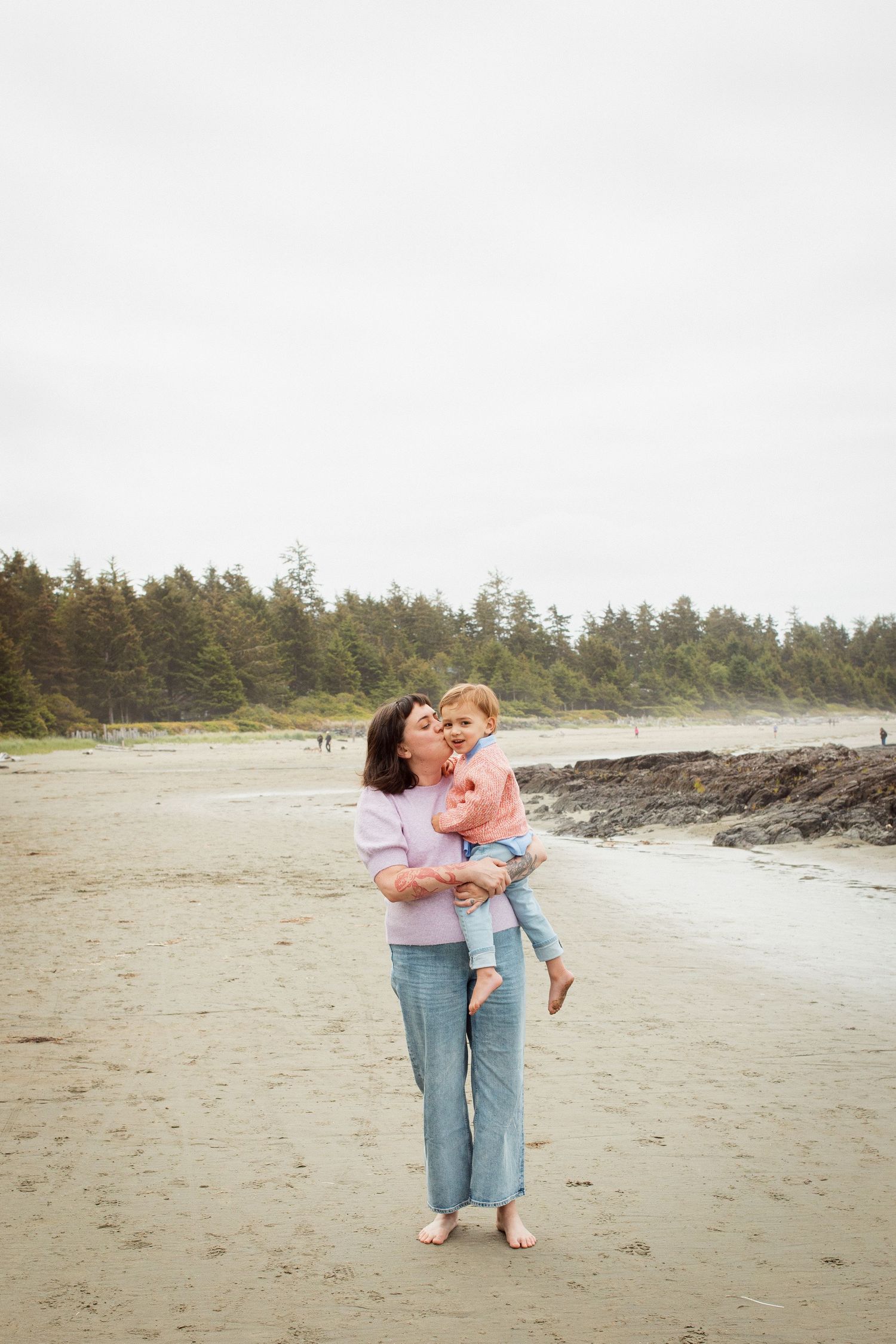 Mother kissing toddler daughter while standing barefoot on sandy beach with forest backdrop.