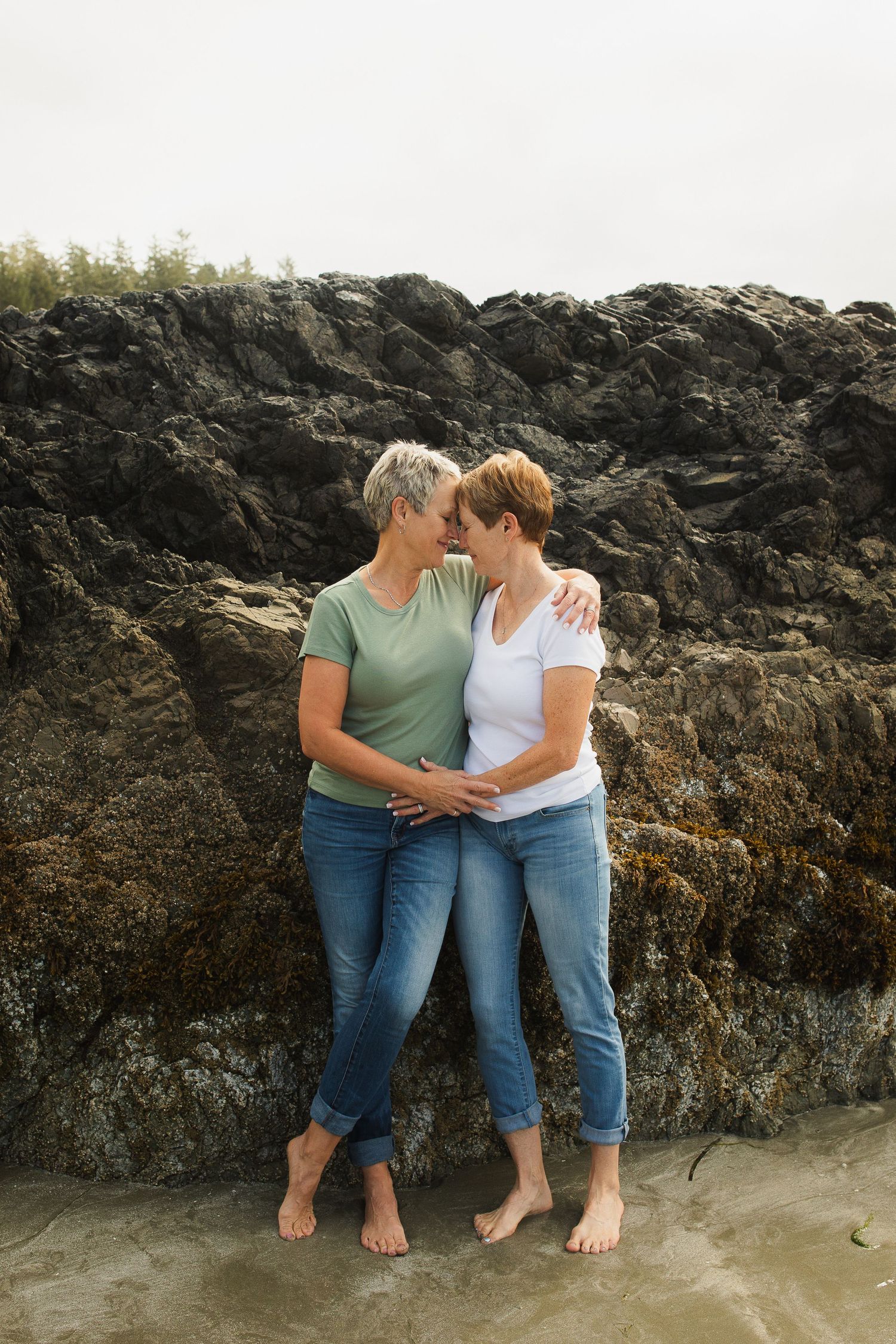 Two barefoot women with short hair share an intimate moment leaning against moss-covered coastal rocks.