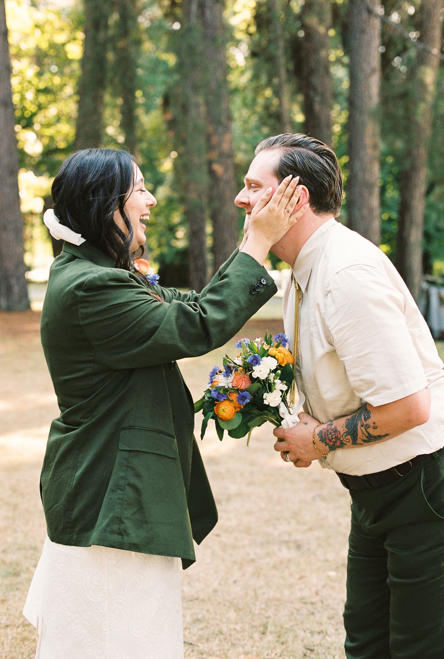 Bride in green jacket laughing while touching grooms face during first look in forest.