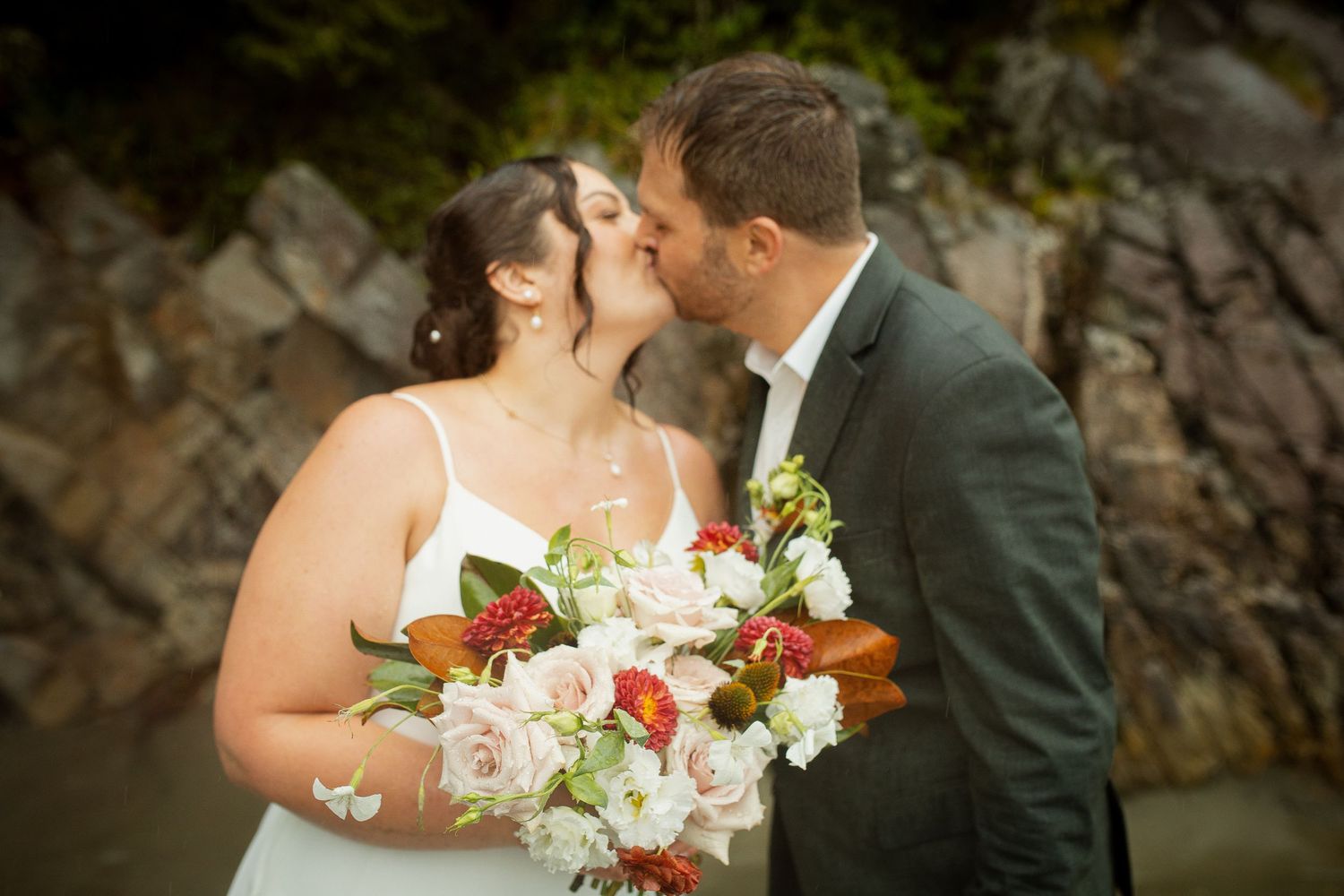 Bride and groom share a romantic kiss outdoors, bride holding a beautiful bouquet of pink and red flowers.