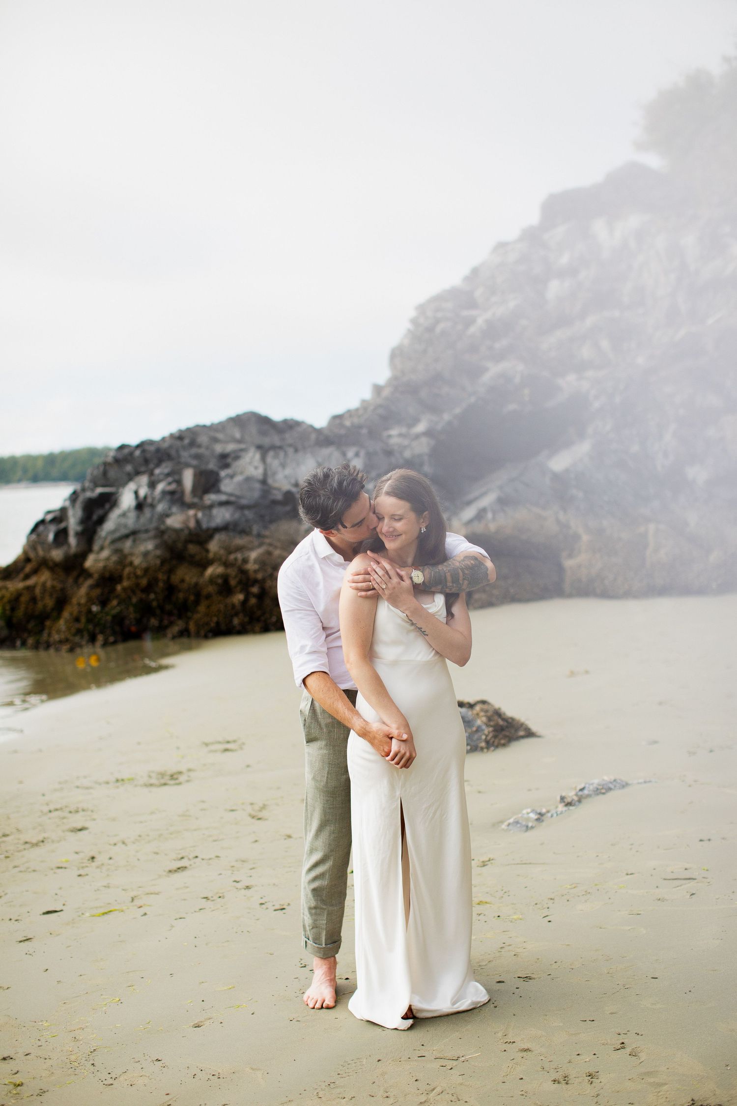 Groom kissing bride on cheek while embracing on a rocky beach.