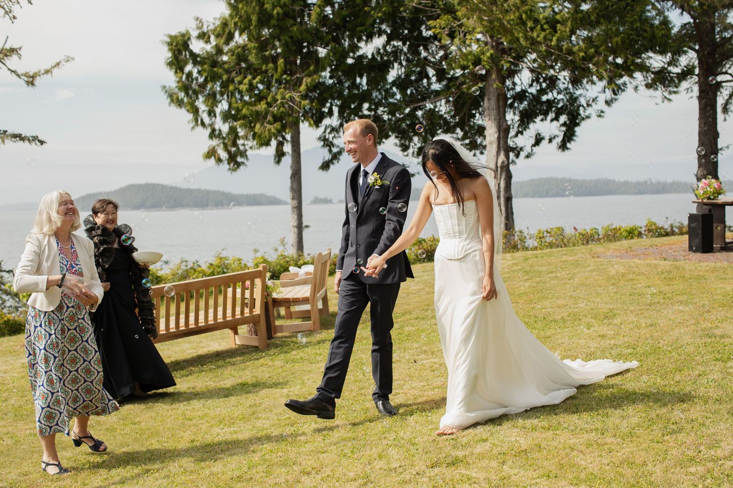 Bride in white gown and groom in navy suit share a first look on a scenic waterfront lawn.
