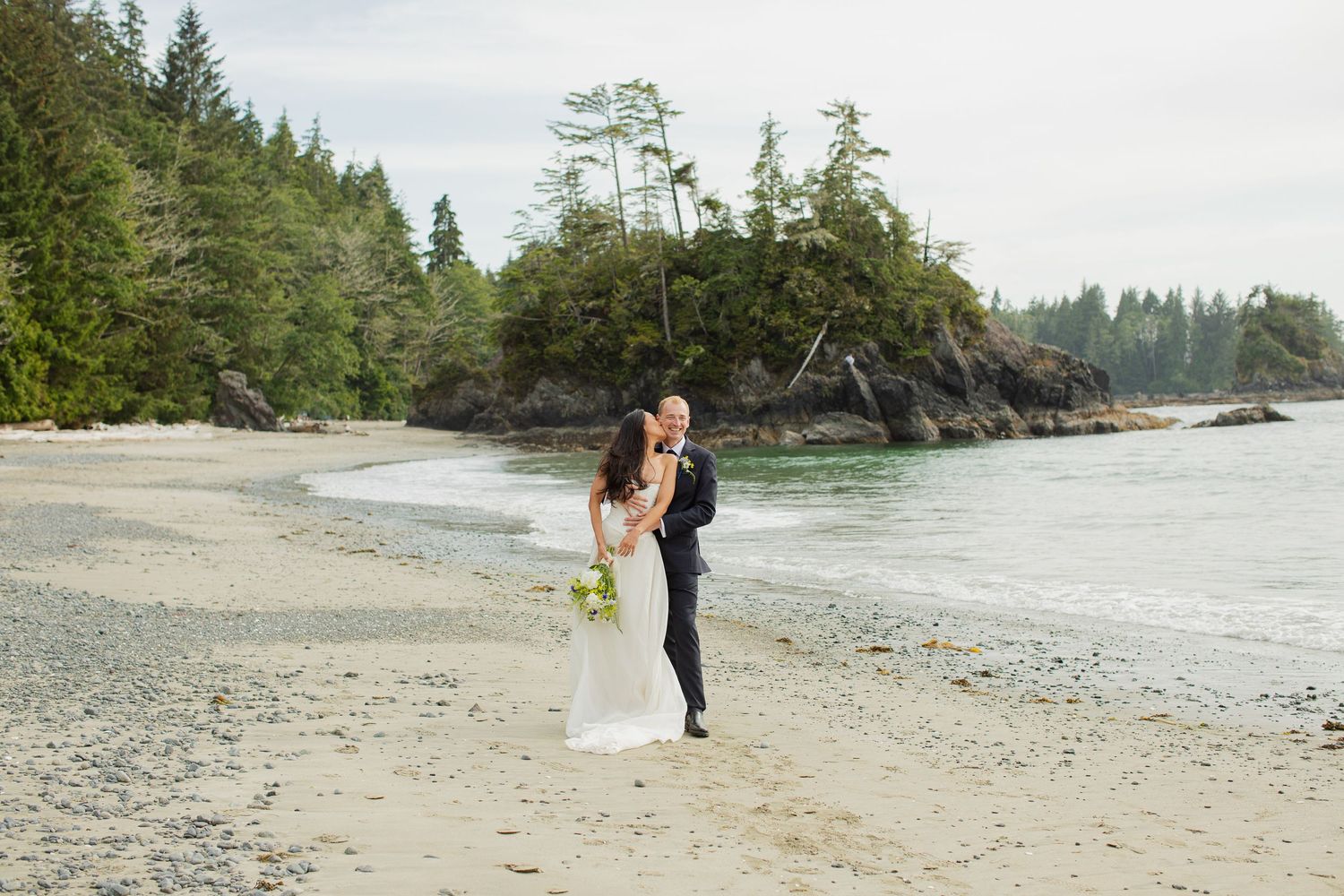 Bride and groom share a kiss on a scenic sandy beach with turquoise water and evergreen trees behind.