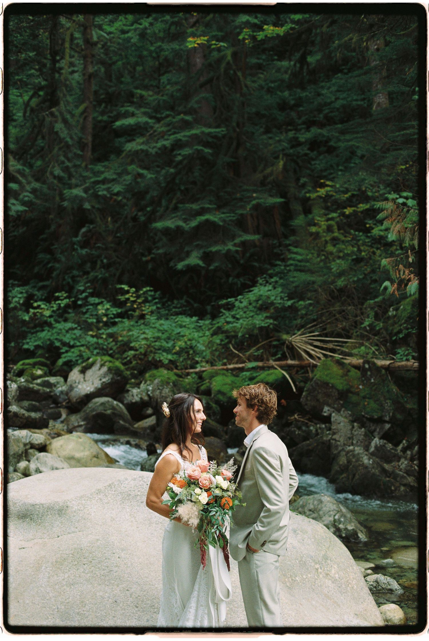 Wedding couple stands on rocky riverbank holding bouquet, surrounded by lush Pacific Northwest forest greenery.