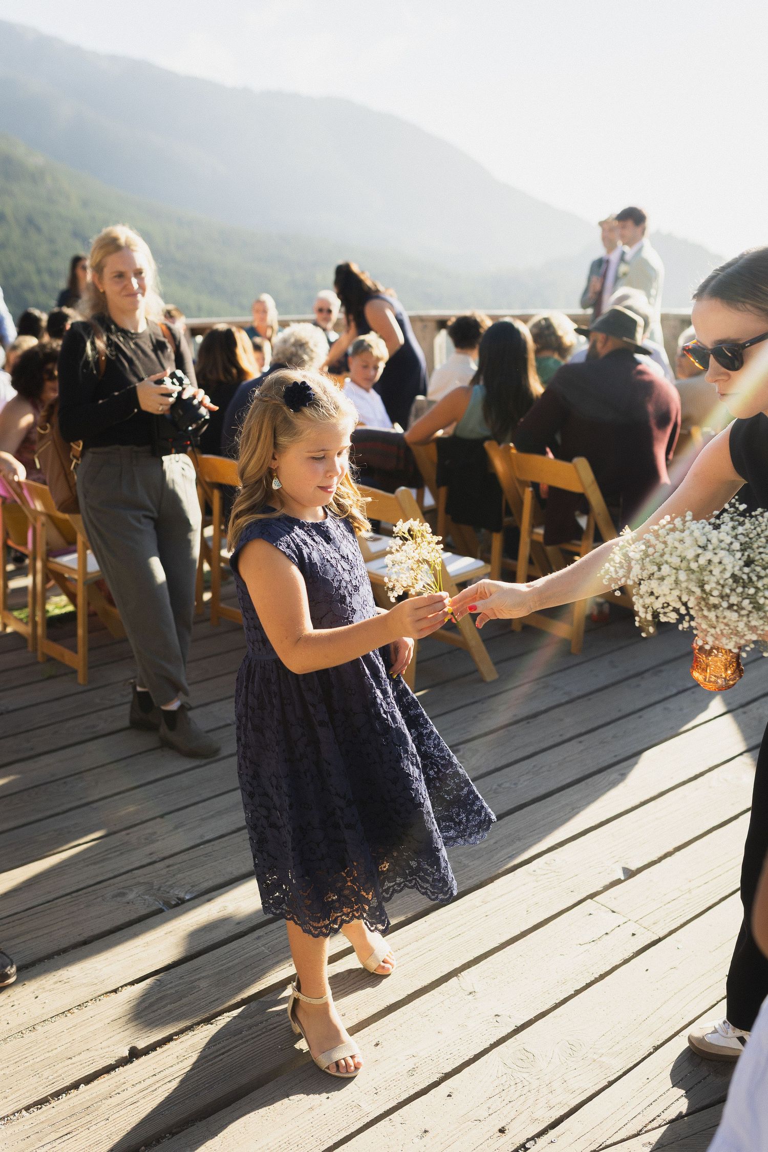 Young flower girl in navy lace dress receives flowers during an outdoor mountain wedding ceremony.