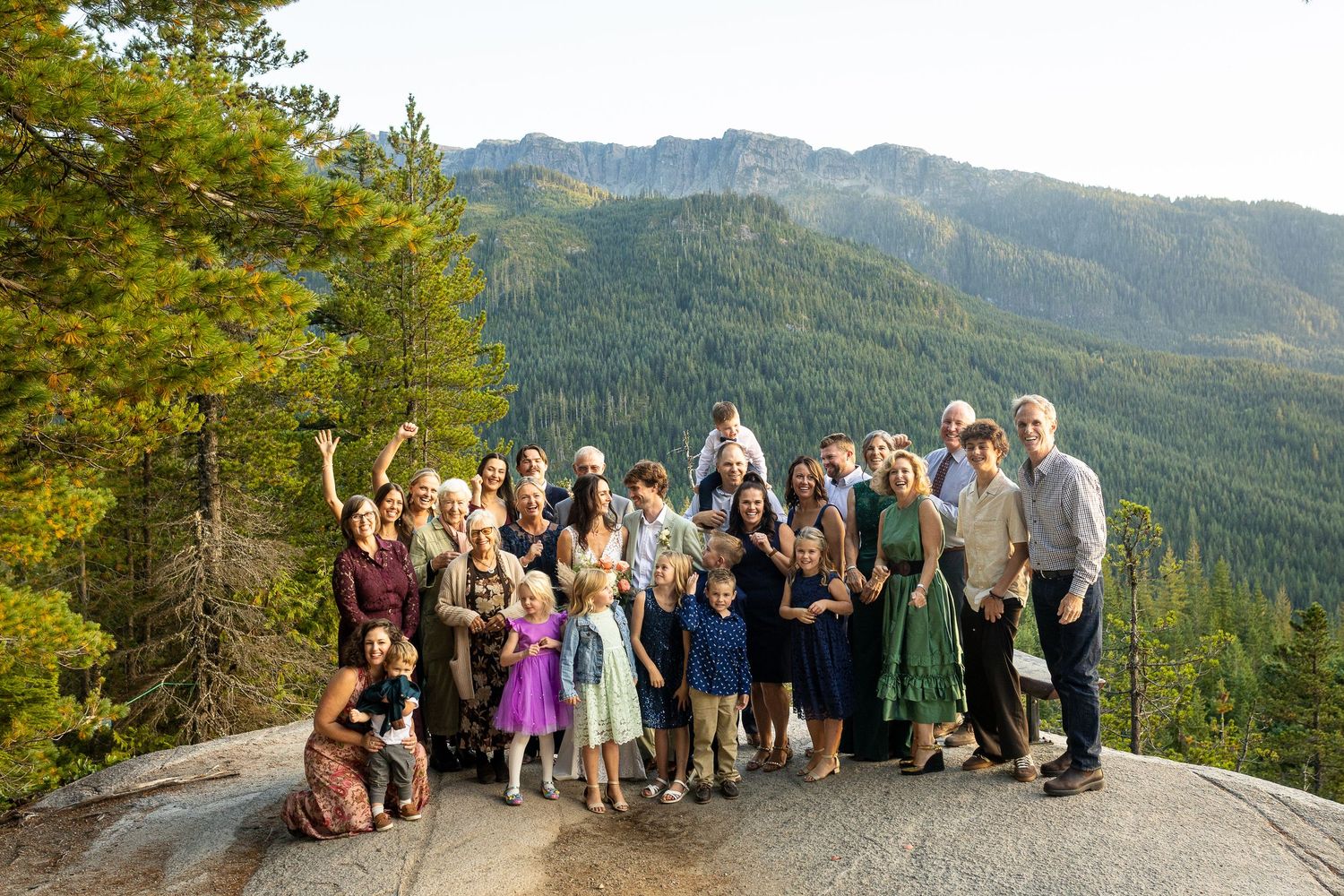 Large wedding party group photo on mountain overlook surrounded by pine trees and scenic peaks.