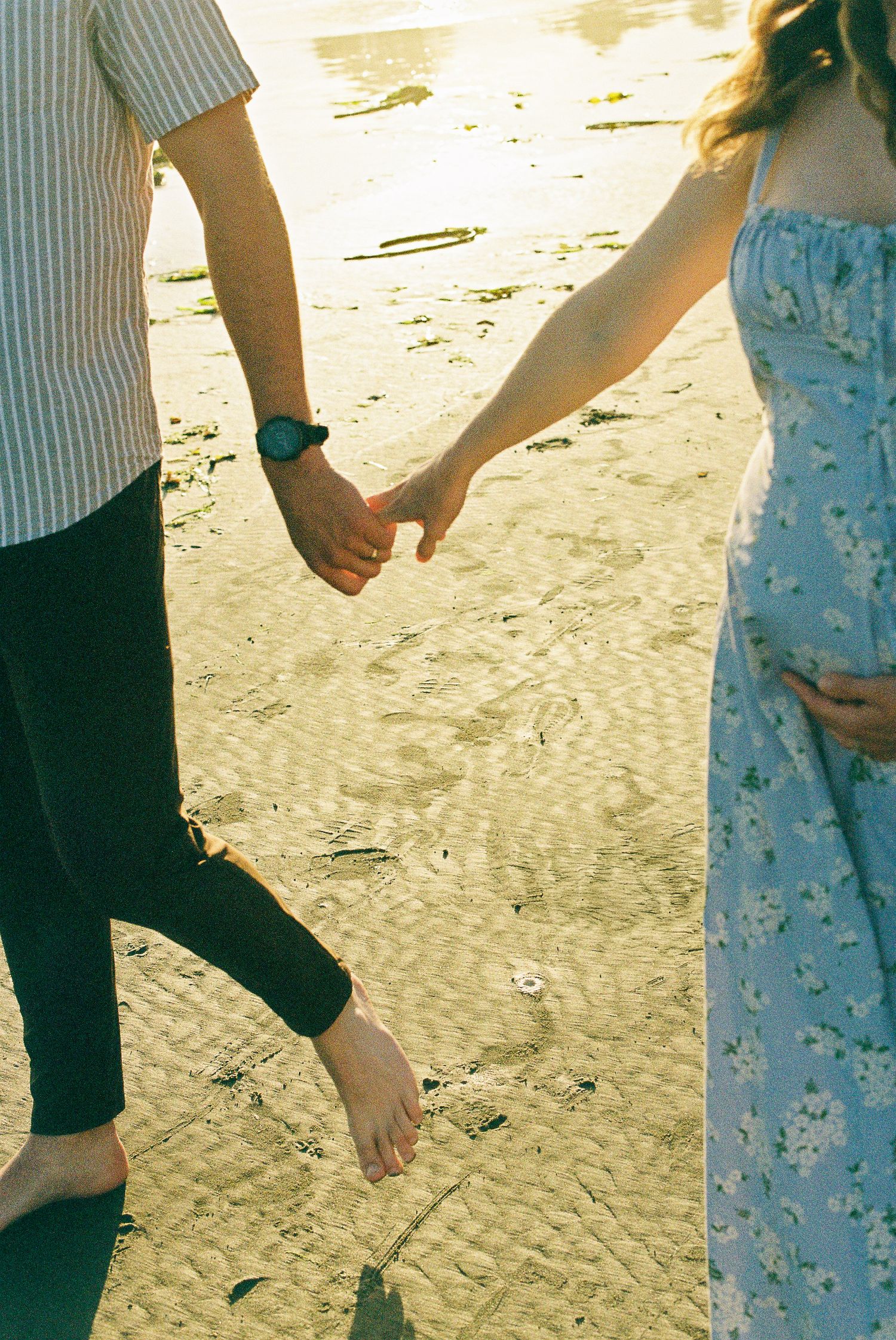 Couple holding hands while walking on a sunny beach, pregnant woman in blue floral dress cradling her bump.
