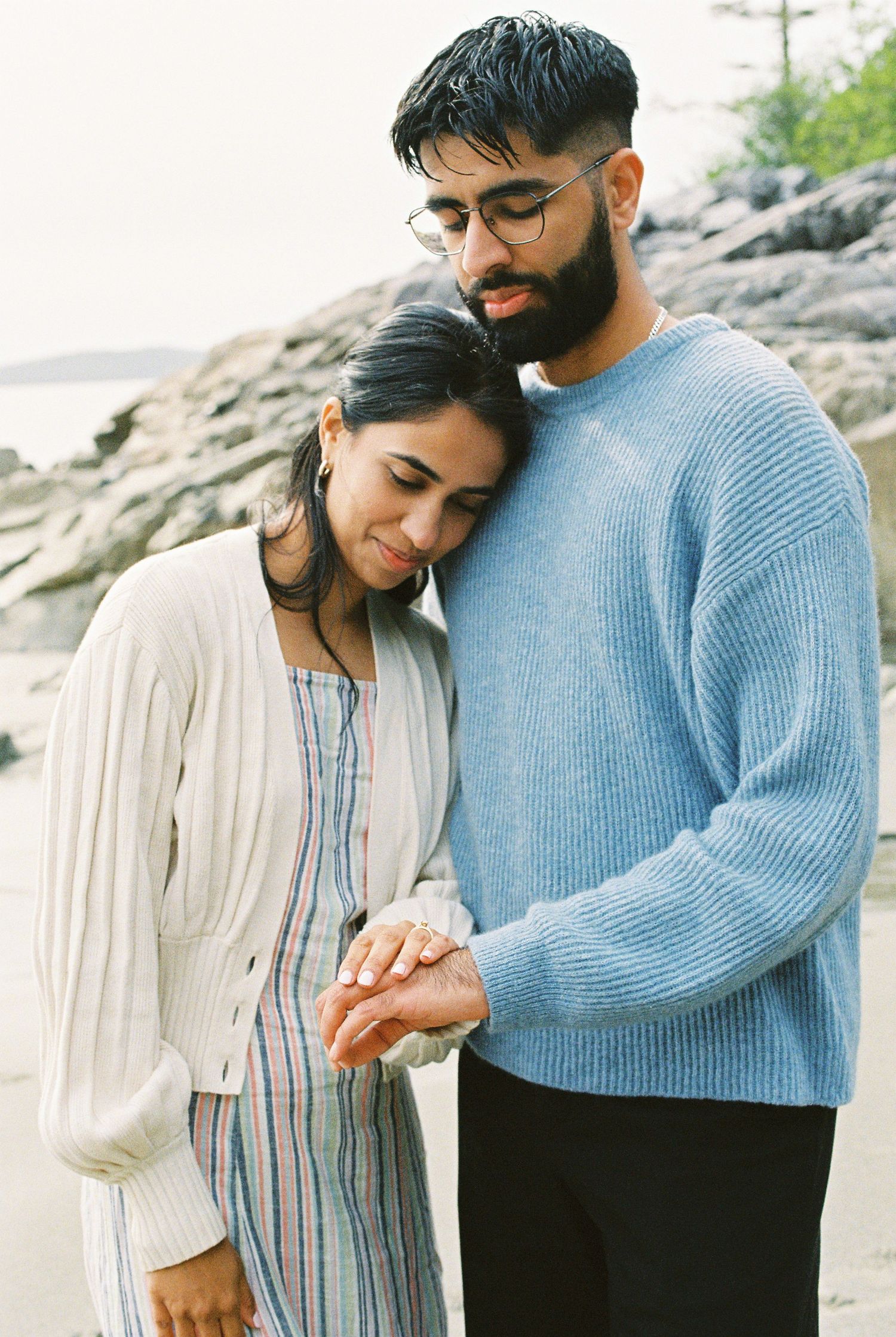 Romantic couple on rocky beach examining engagement ring, man in blue sweater and woman in white cardigan with striped dress.