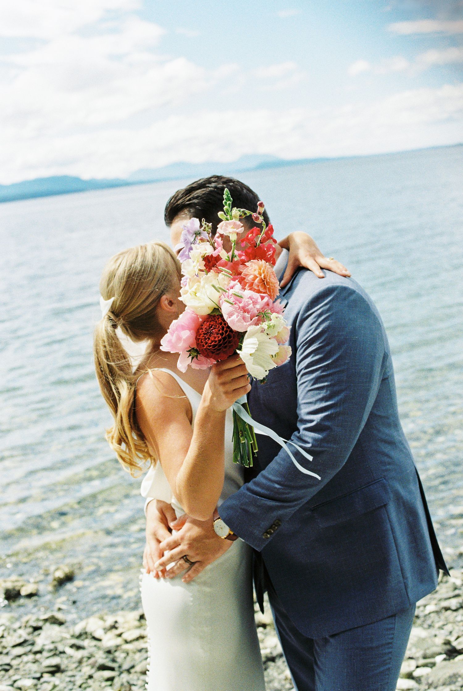 Bride and groom sharing a kiss behind a vibrant colorful bouquet by a lakeside shore.