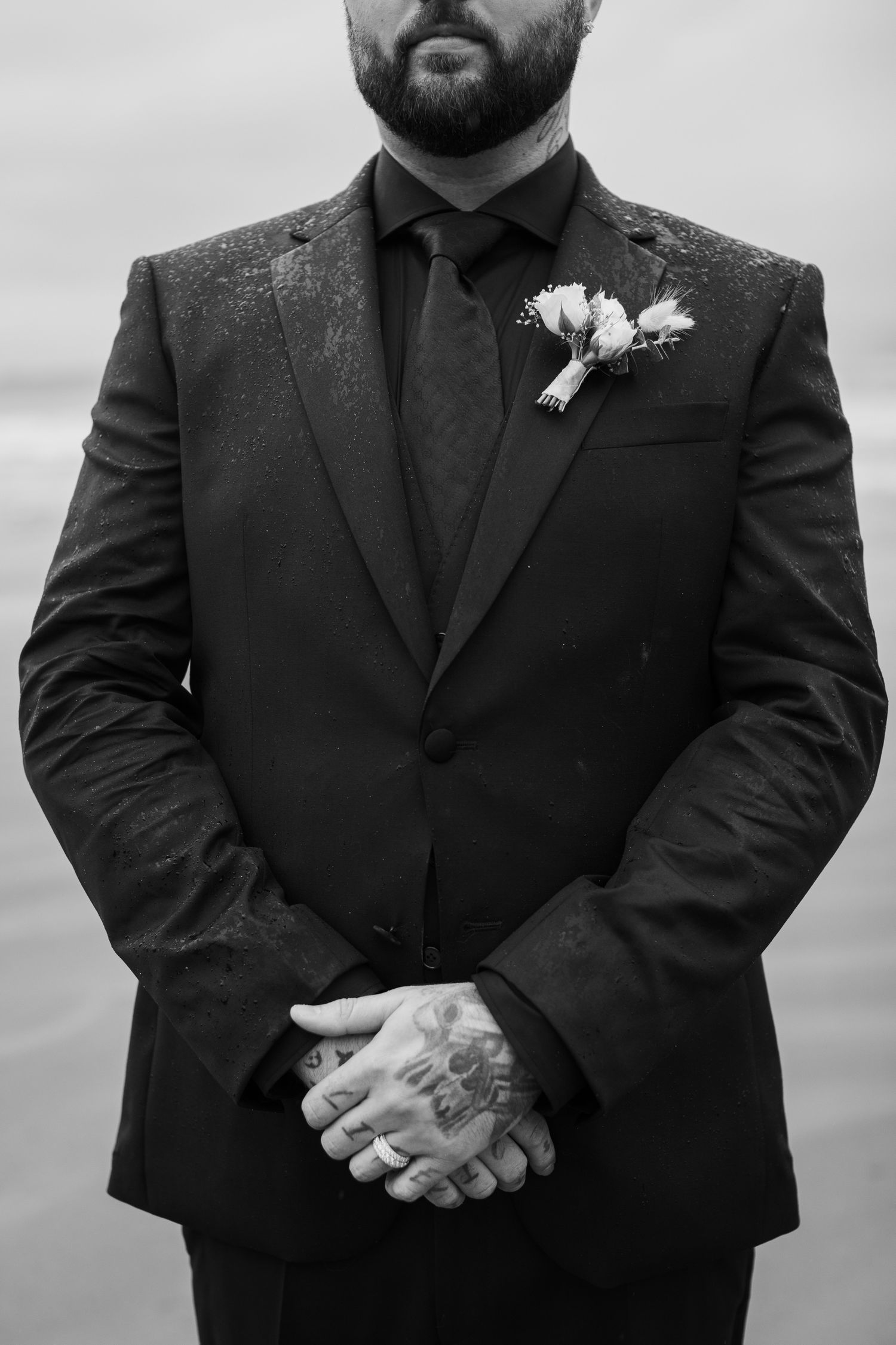 Black and white close-up of tattooed groom in dark suit and tie holding hands at wedding.
