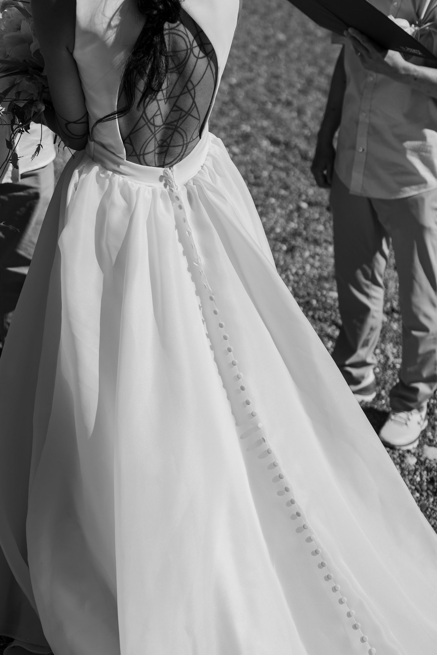 Black and white close-up of bride's elegant backless wedding gown with delicate lace detail and button closure.