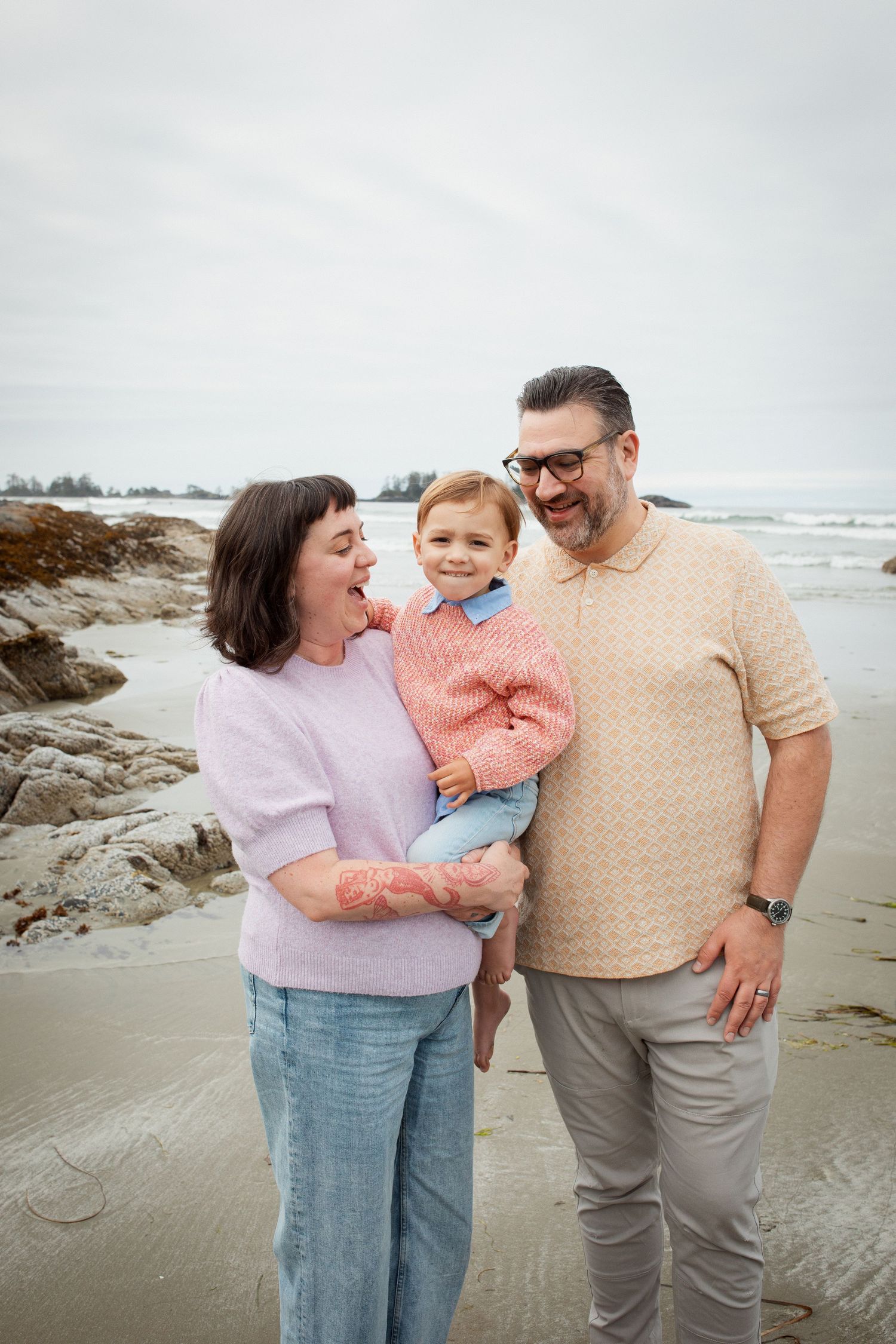 Two parents smiling with young child on overcast beach with ocean waves in background.