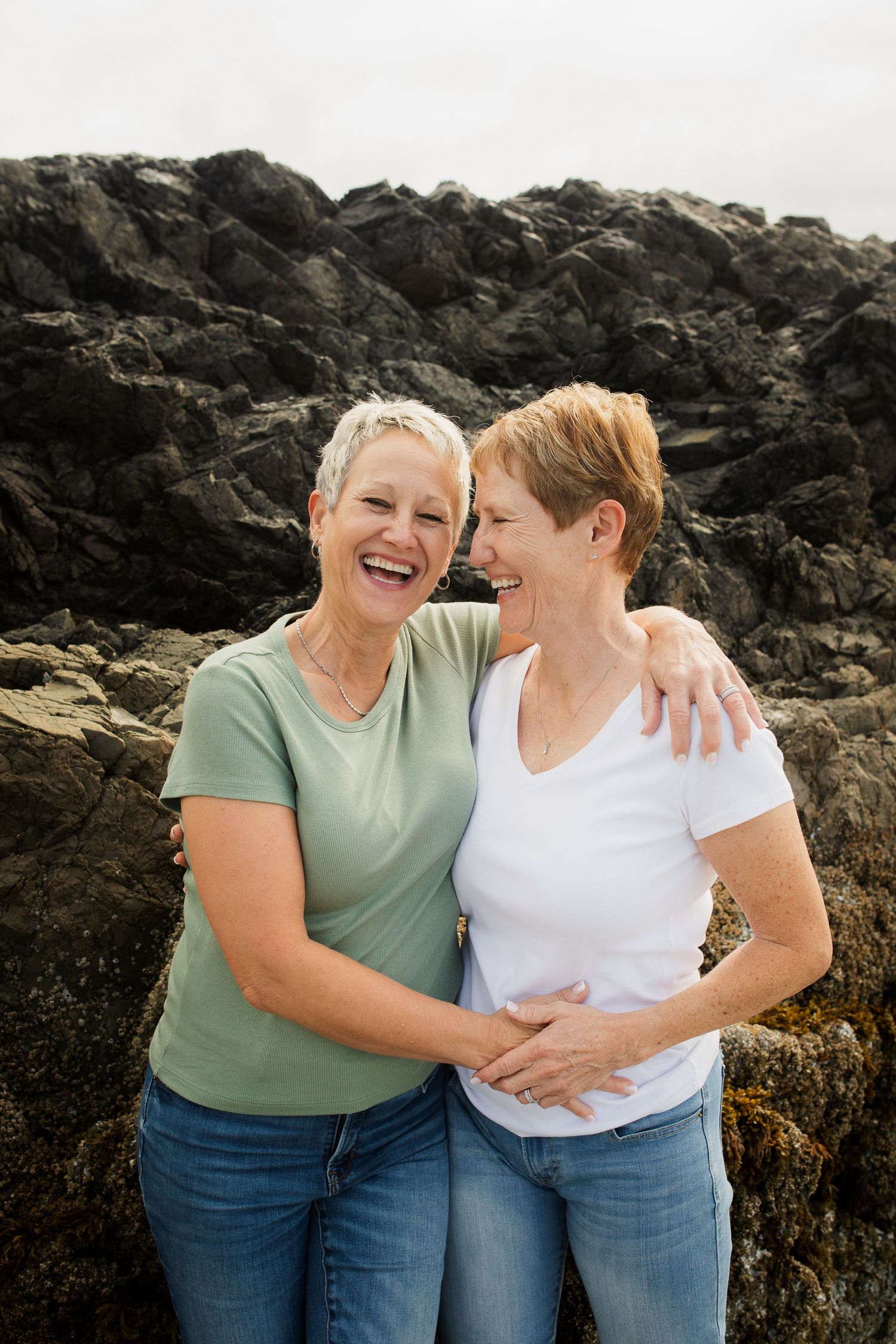 Two mature women with short hair laugh joyfully together while embracing in front of rocky coastal cliffs.