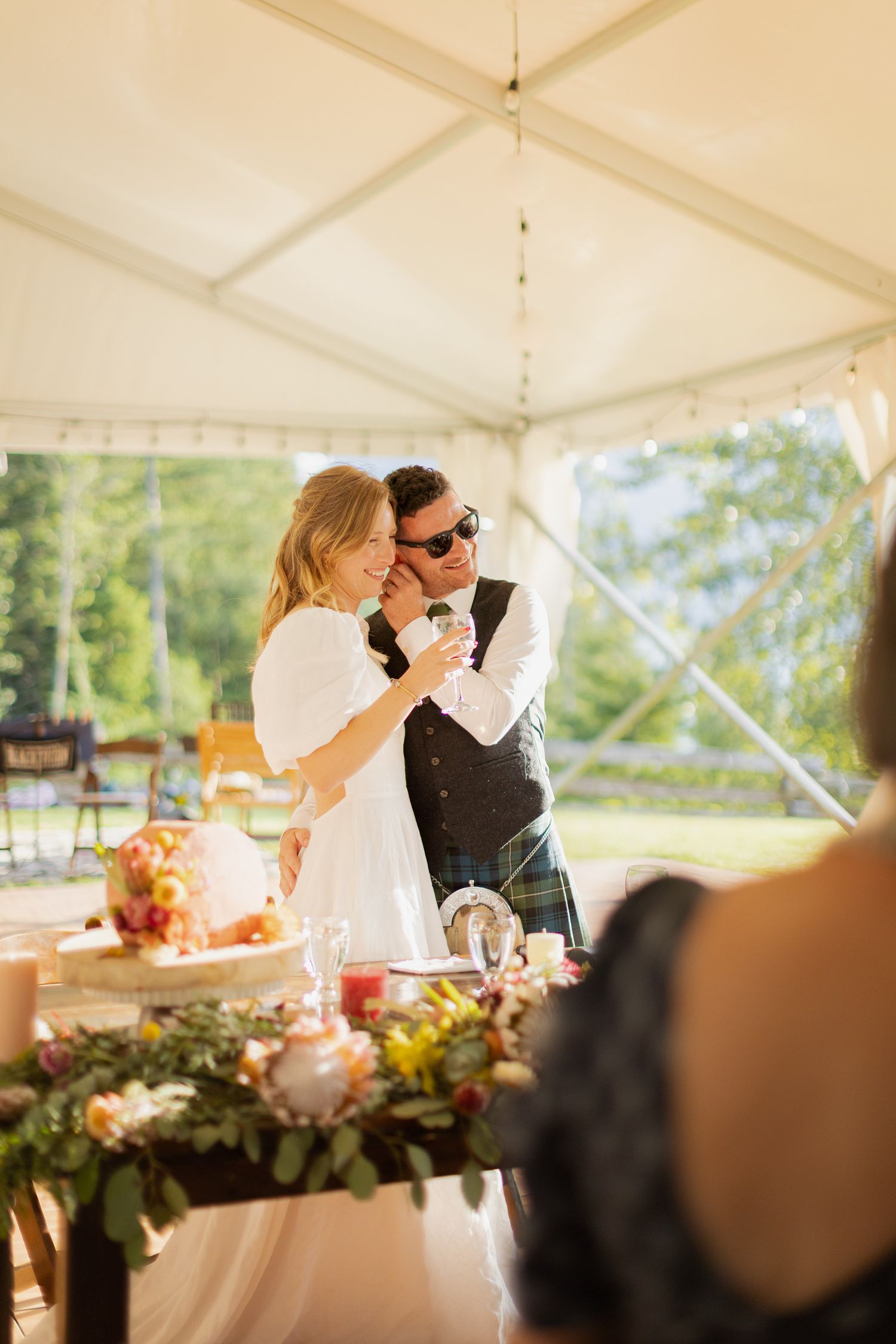 Bride and groom share first dance under white tent at outdoor wedding reception with floral table decorations.