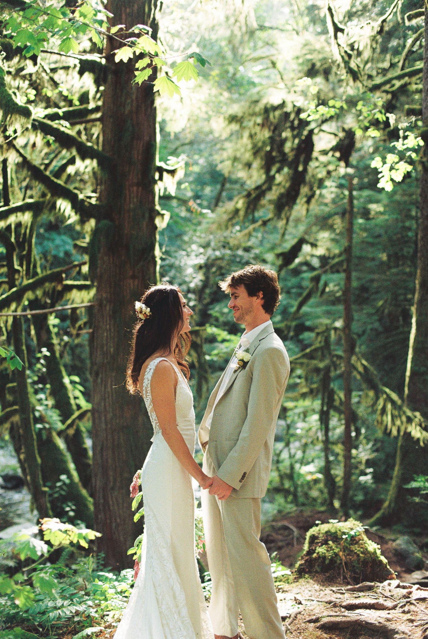 Bride and groom holding hands and gazing at each other in sunlit mossy forest setting.
