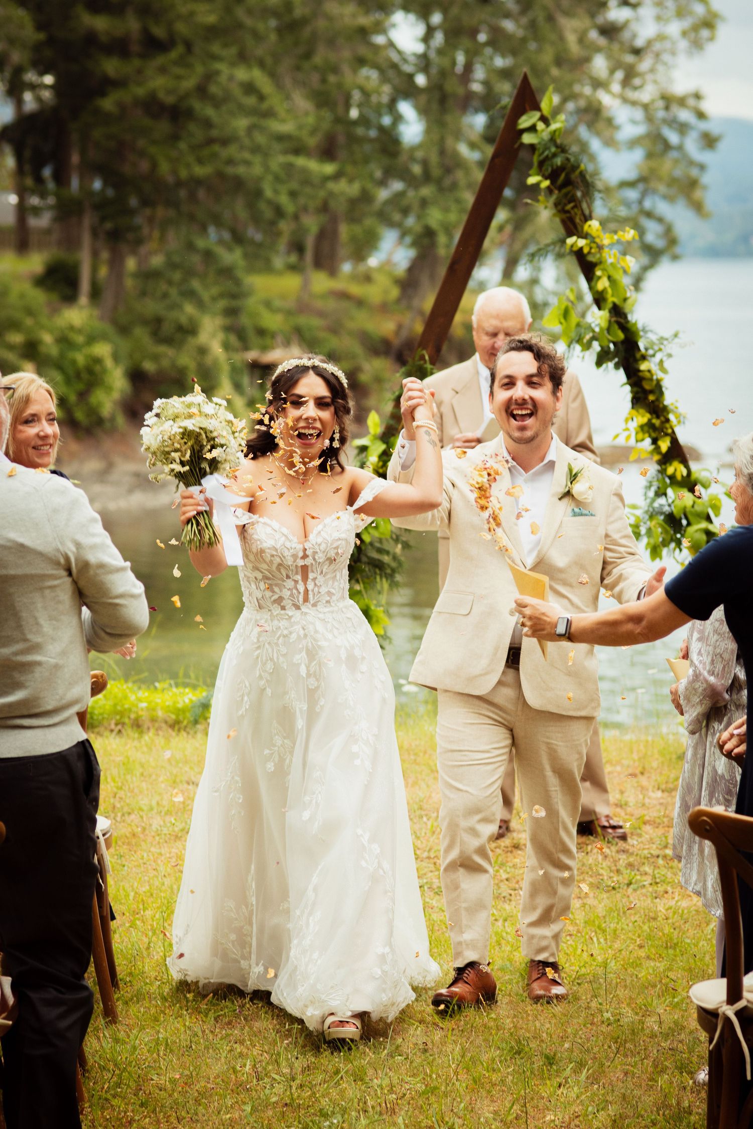 Joyful bride and groom walking down aisle as guests toss confetti at outdoor lakeside wedding.