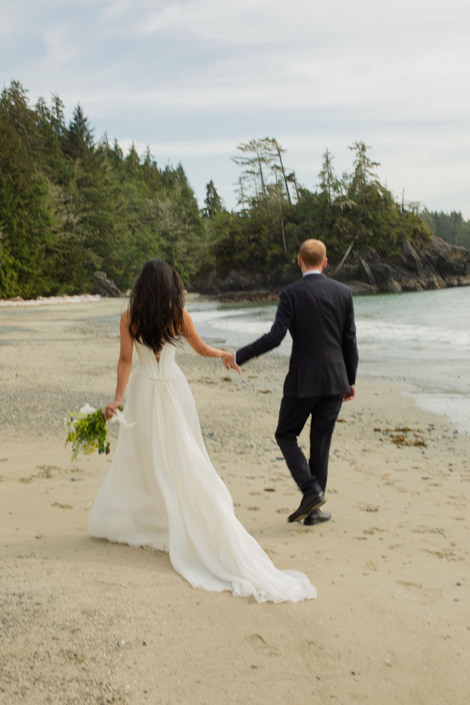 Bride and groom walk hand-in-hand along sandy beach, bride carrying white bouquet, lush forest backdrop.
