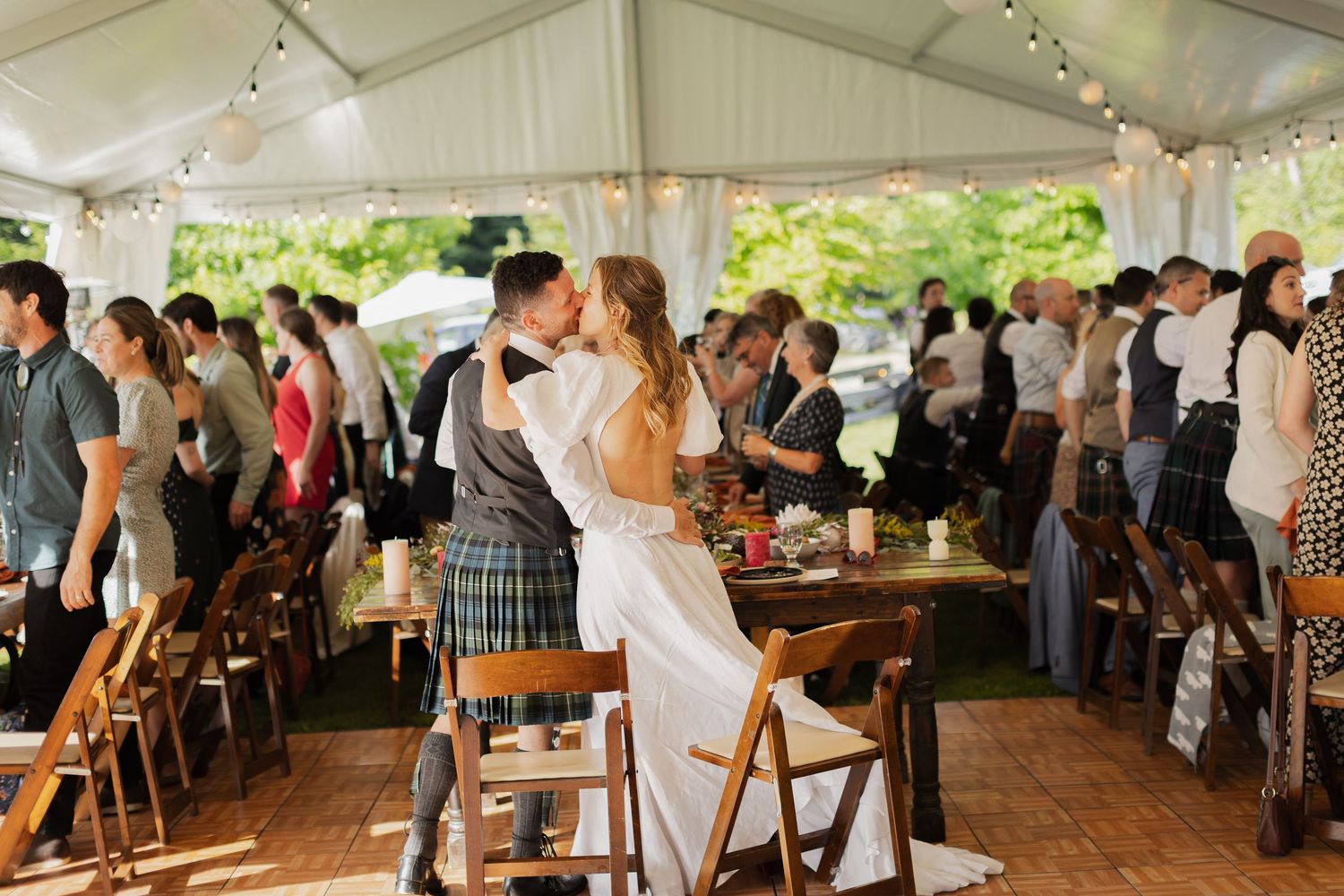 Bride and groom share first dance under white tent at elegant outdoor wedding reception with guests gathered around.