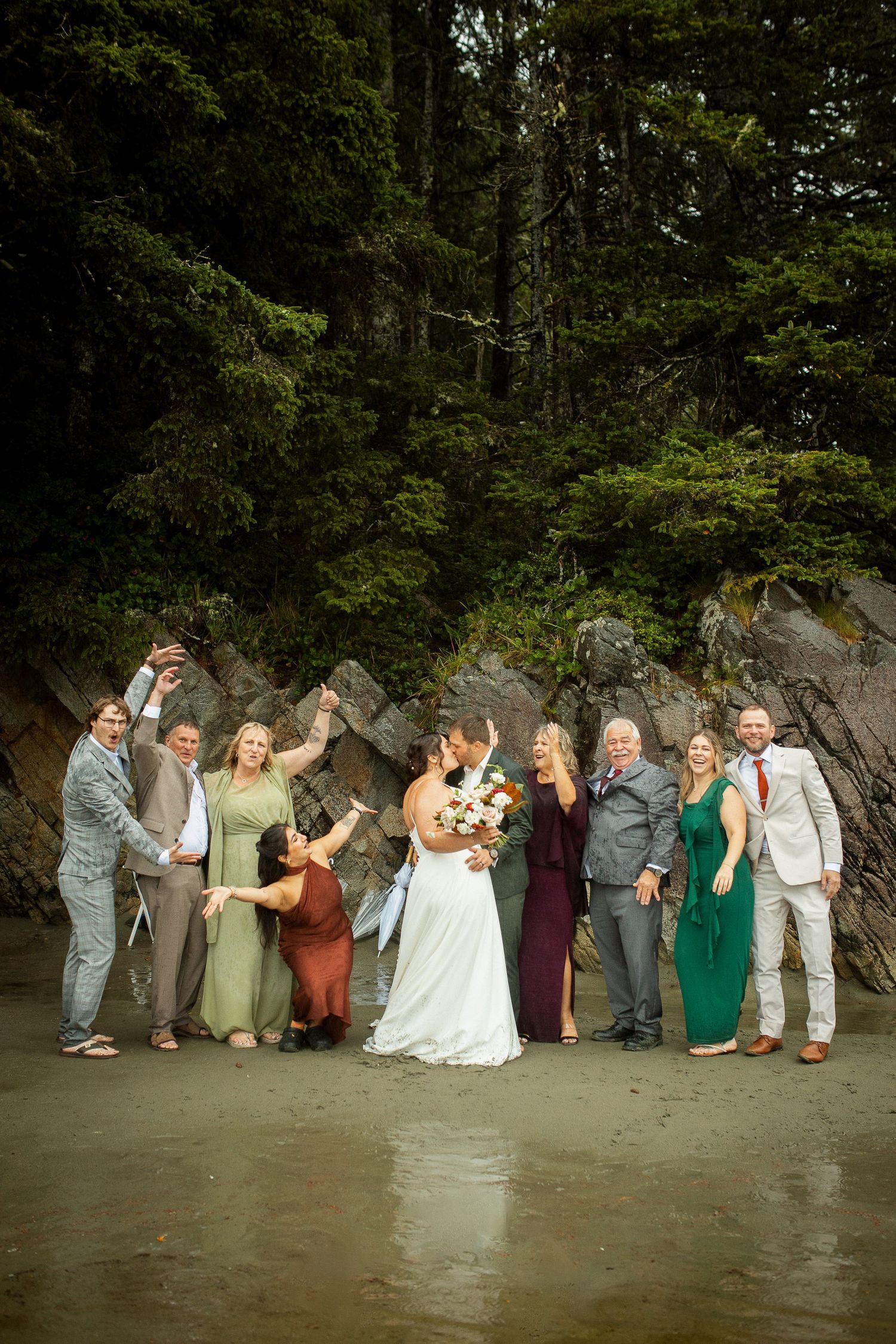 Wedding party poses joyfully on a beach surrounded by lush green forest and rocky cliffs.