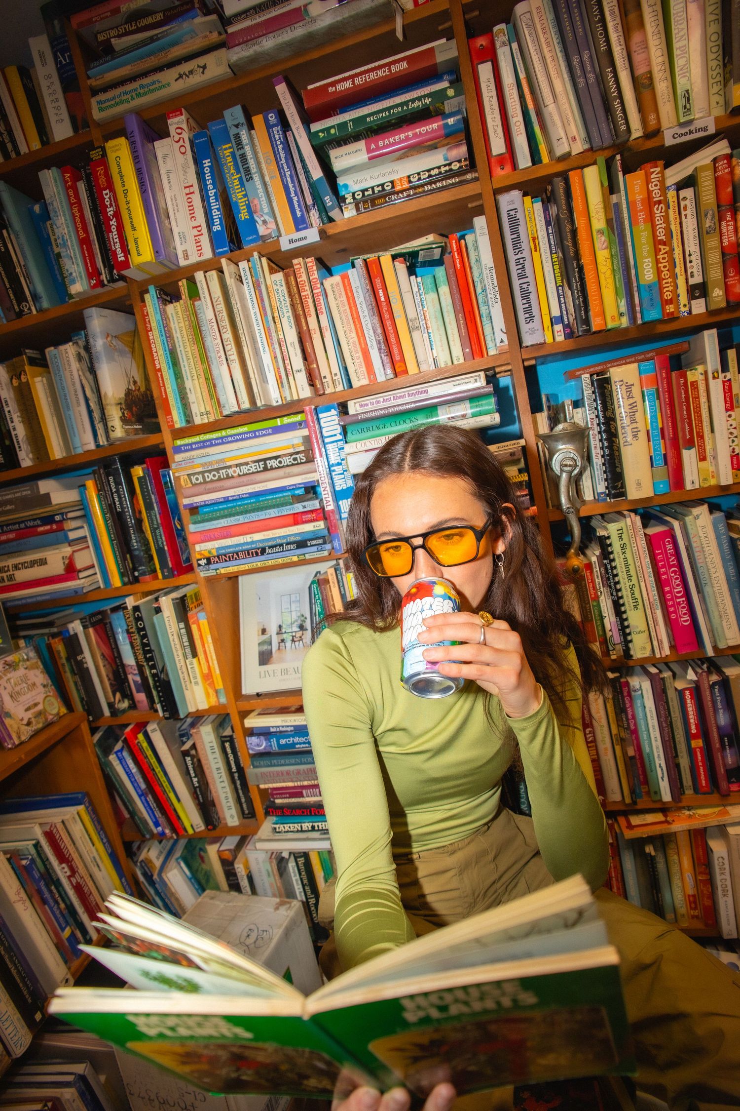 Young woman with orange tinted glasses reading book and drinking can in colorful bookstore.
