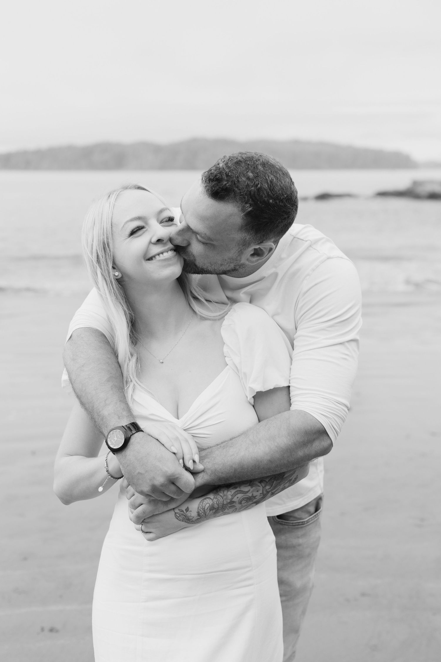 Black and white photo of a couple embracing on a beach, man kissing woman's cheek as she smiles.