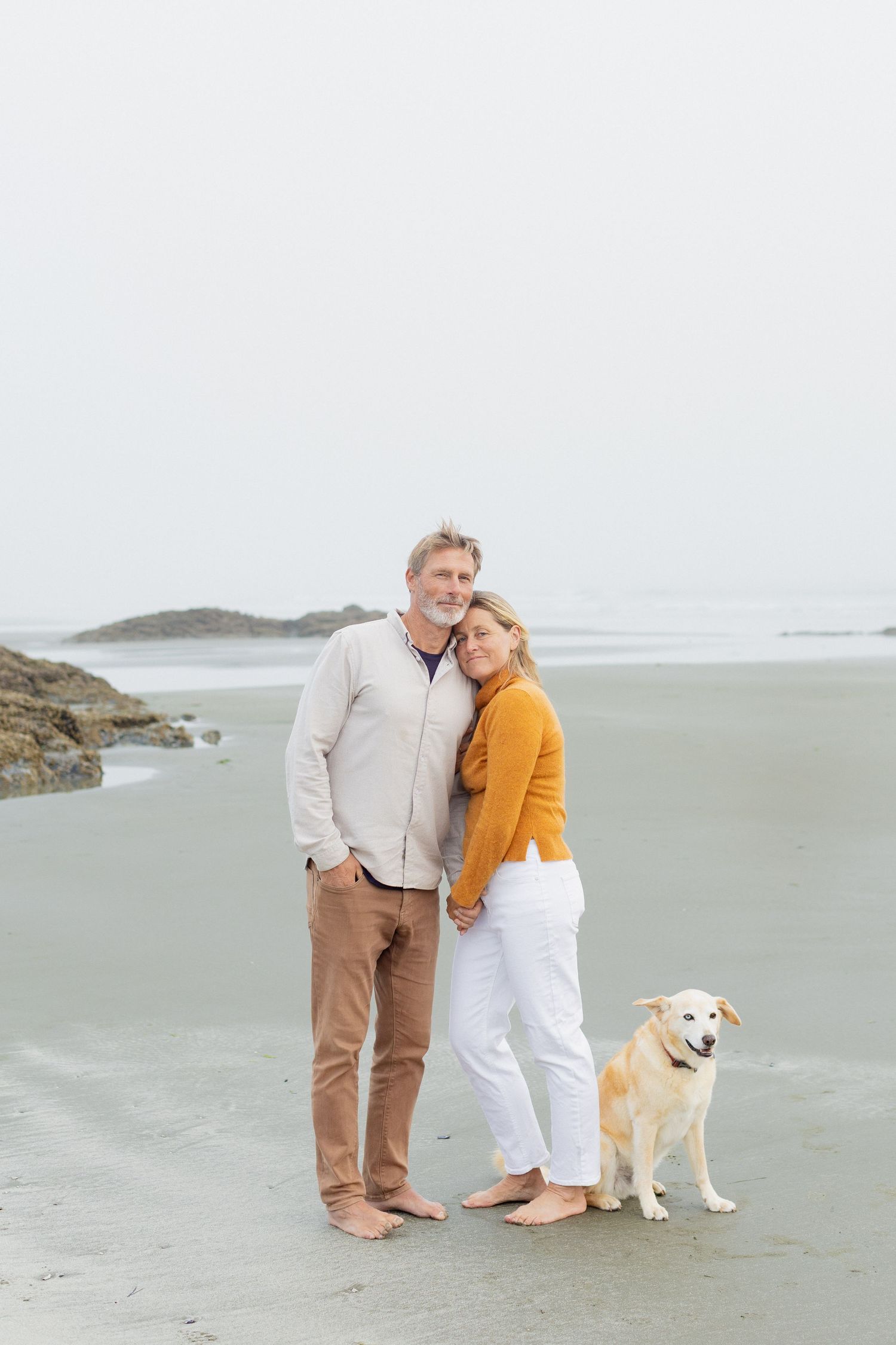 Couple in orange and beige outfits posing with golden retriever on overcast foggy beach.