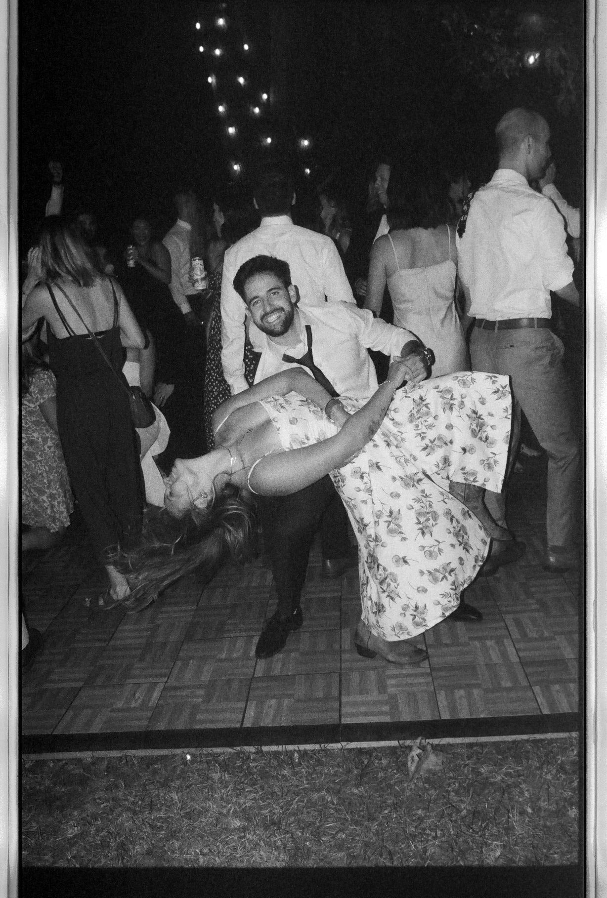 Black and white photo of groom dipping partner in floral dress on crowded outdoor wedding dance floor.
