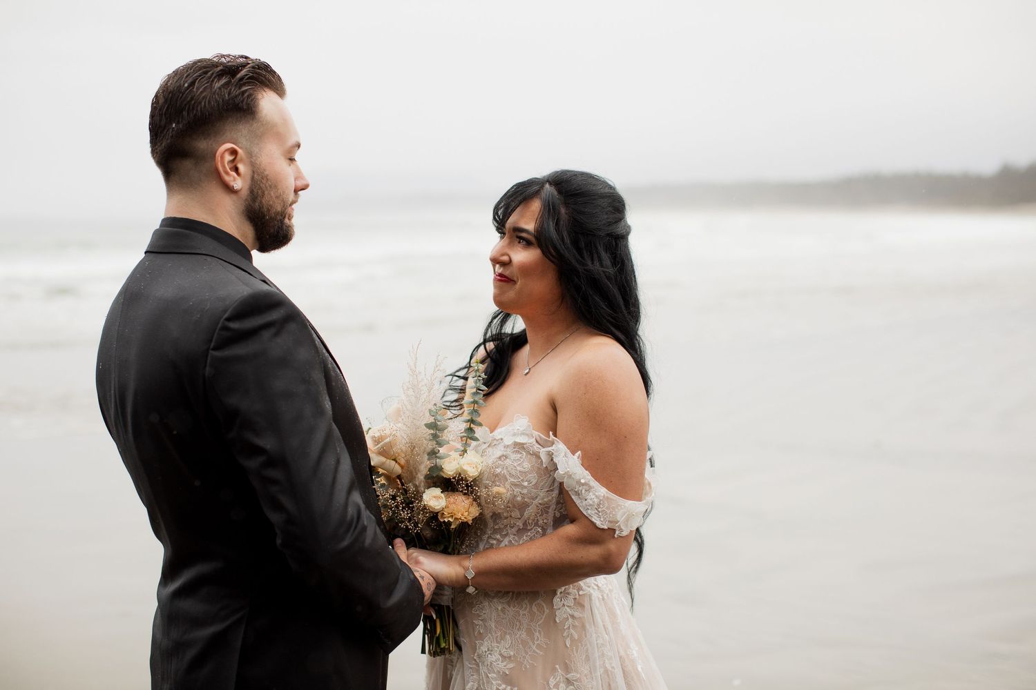 Bride and groom exchanging vows on overcast beach, bride holding dried bouquet.