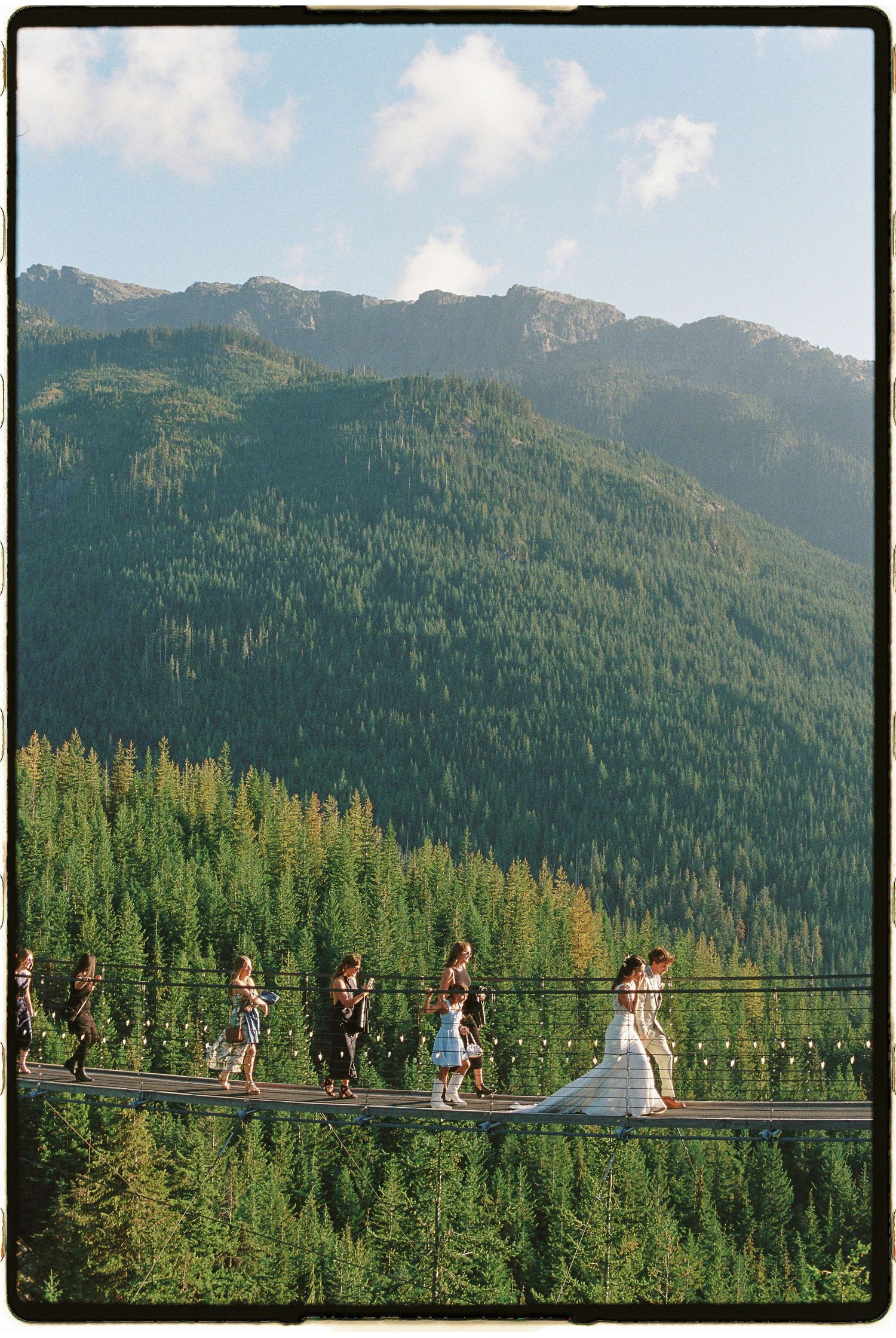 Wedding party crosses a scenic suspension bridge surrounded by lush green mountain forest on a sunny day.