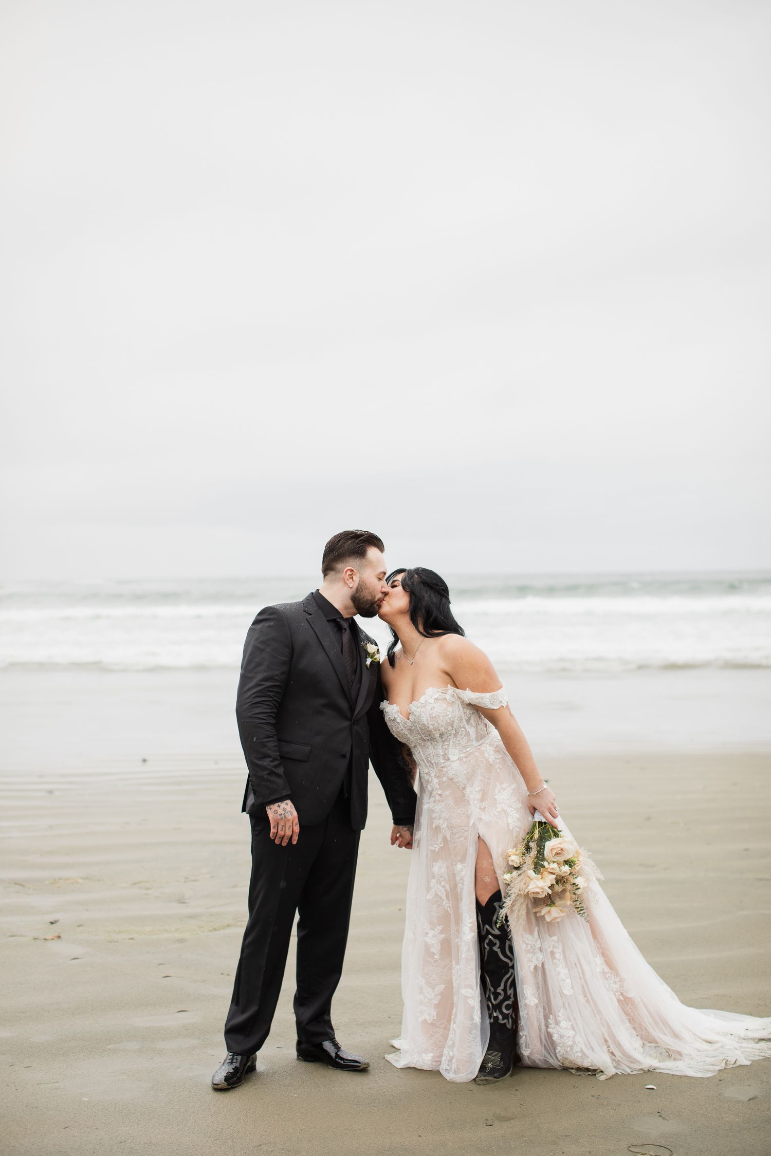 Newlyweds share a romantic kiss on a foggy beach, bride holding bouquet in lace gown with train.