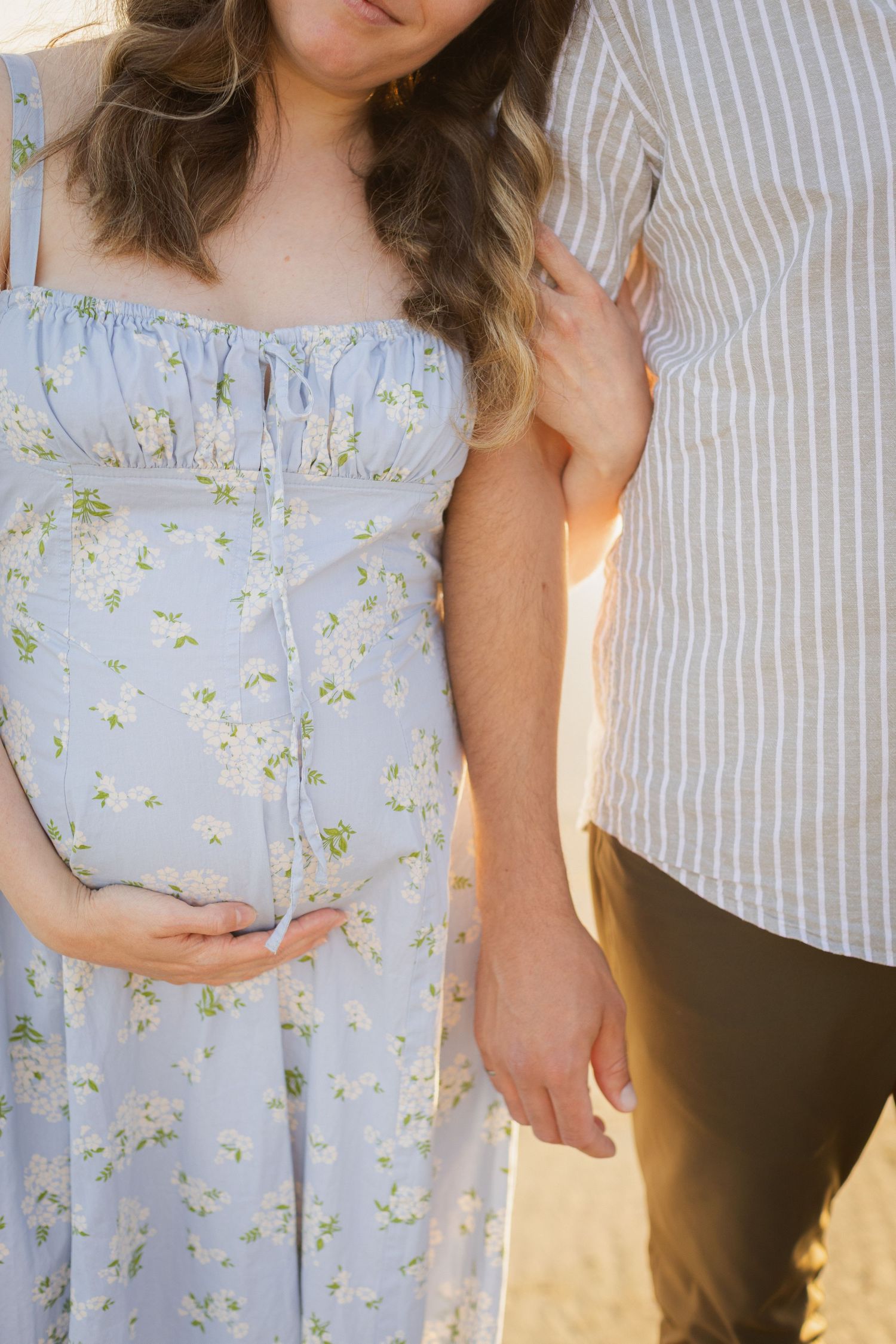 Close-up of pregnant woman in blue floral dress cradling baby bump while partner stands beside her on beach.
