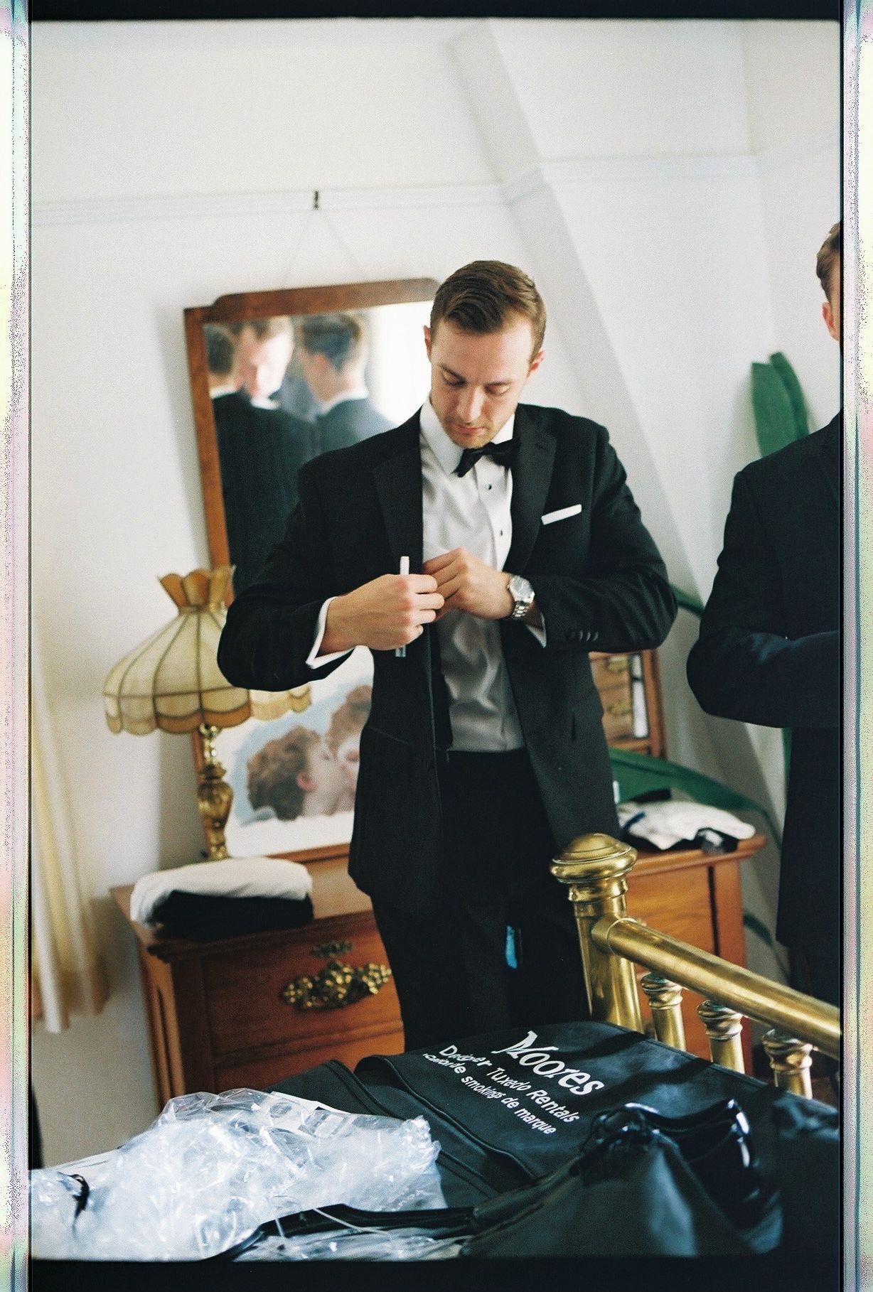 Groom in classic black tuxedo adjusts his shirt in a vintage bedroom mirror before the wedding ceremony.
