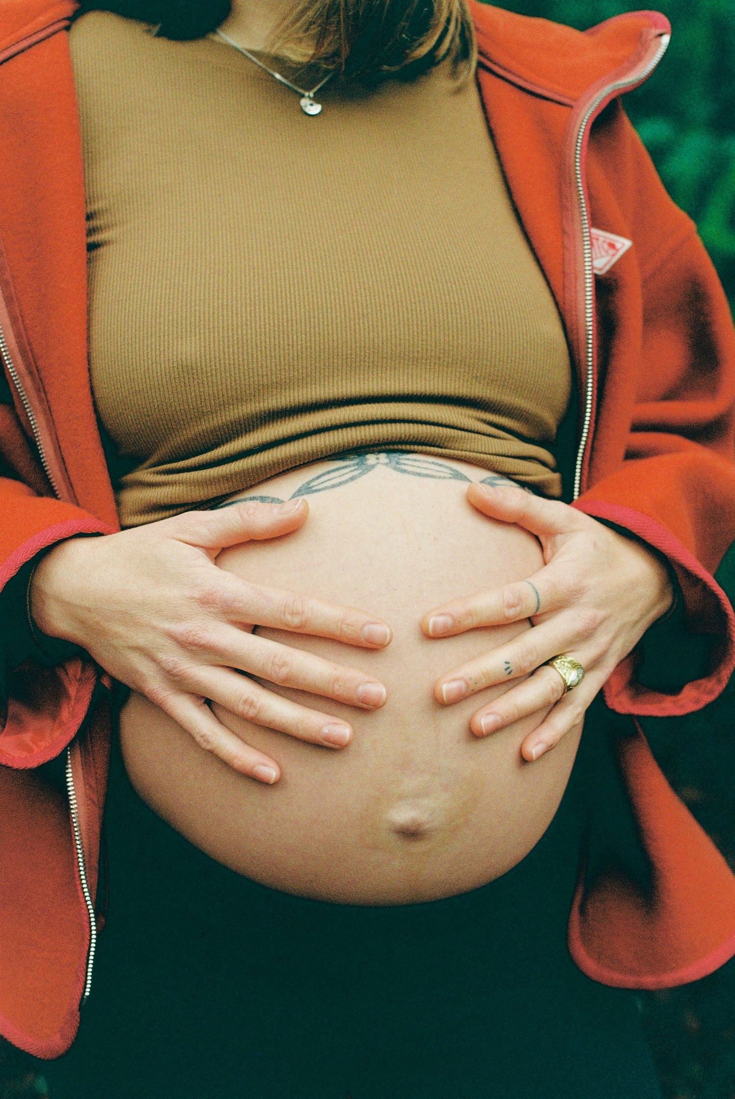 Close-up of pregnant belly held by hands, wearing red jacket and olive top with tattoo visible.