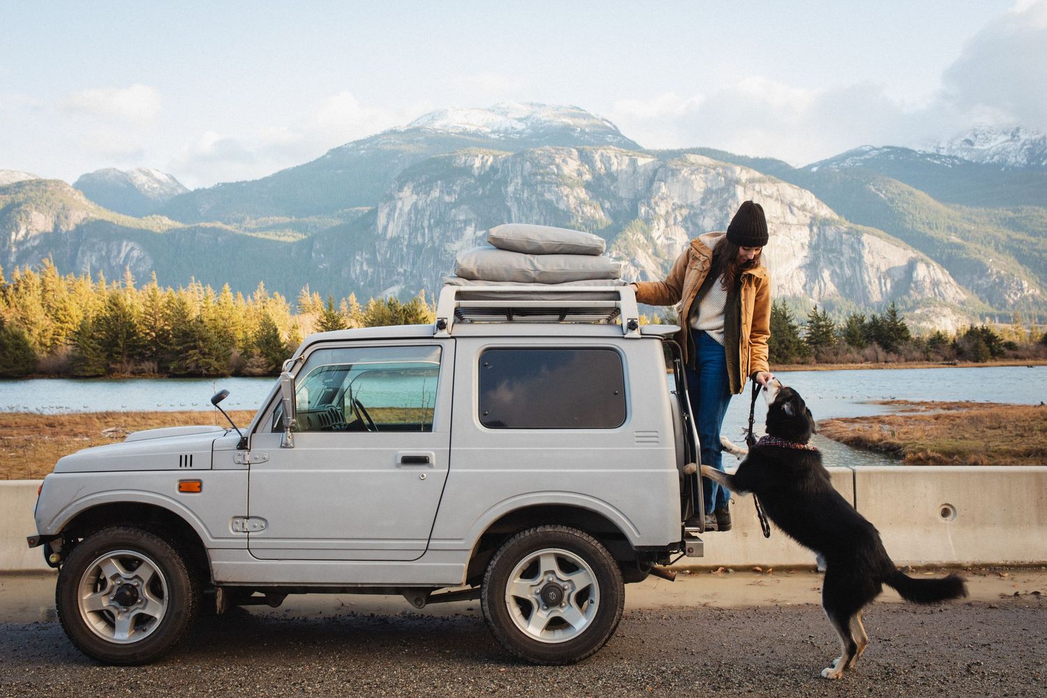 Woman loading gear onto a white Suzuki Jimny roof rack with a black dog jumping up beside her.