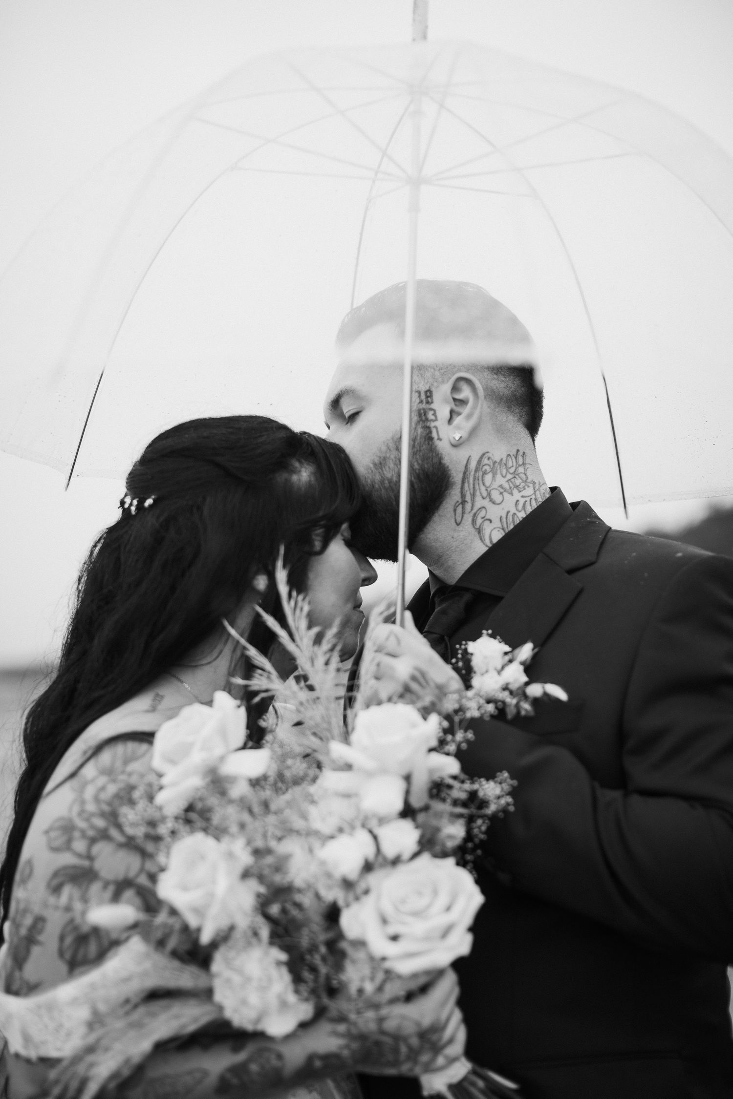 Black and white photo of bride and groom nearly kissing under clear umbrella, bride holding floral bouquet.